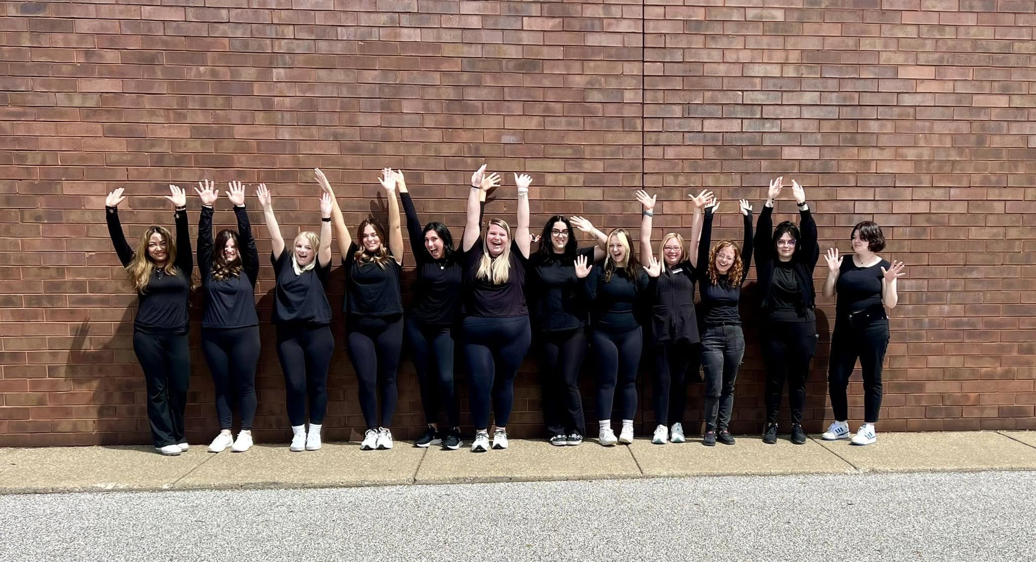 Group of eleven women standing against a brick wall, smiling, with arms raised in celebration.