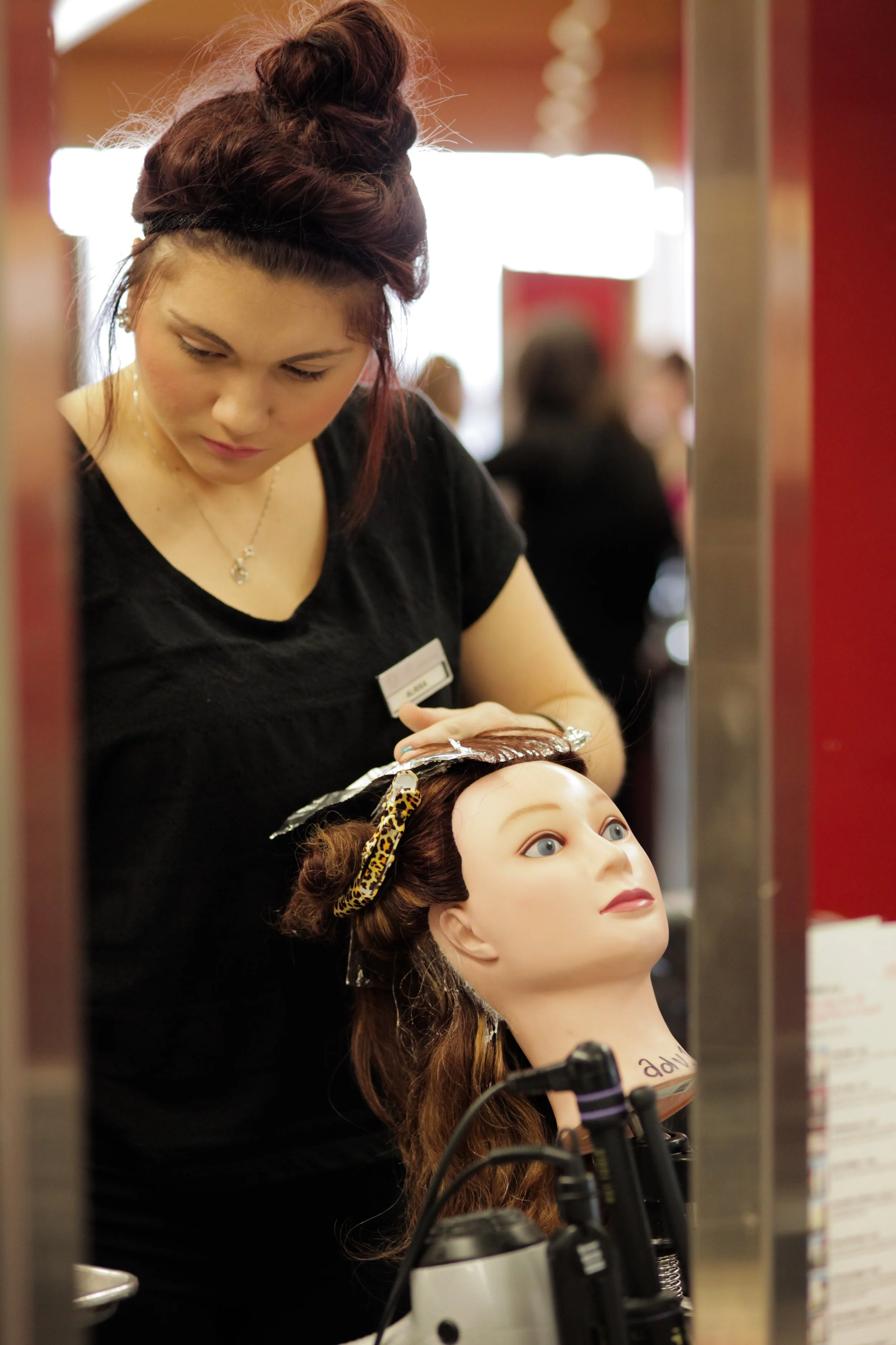 A woman with braided hair and a name tag styling a mannequin's long curly hair at a hair salon.