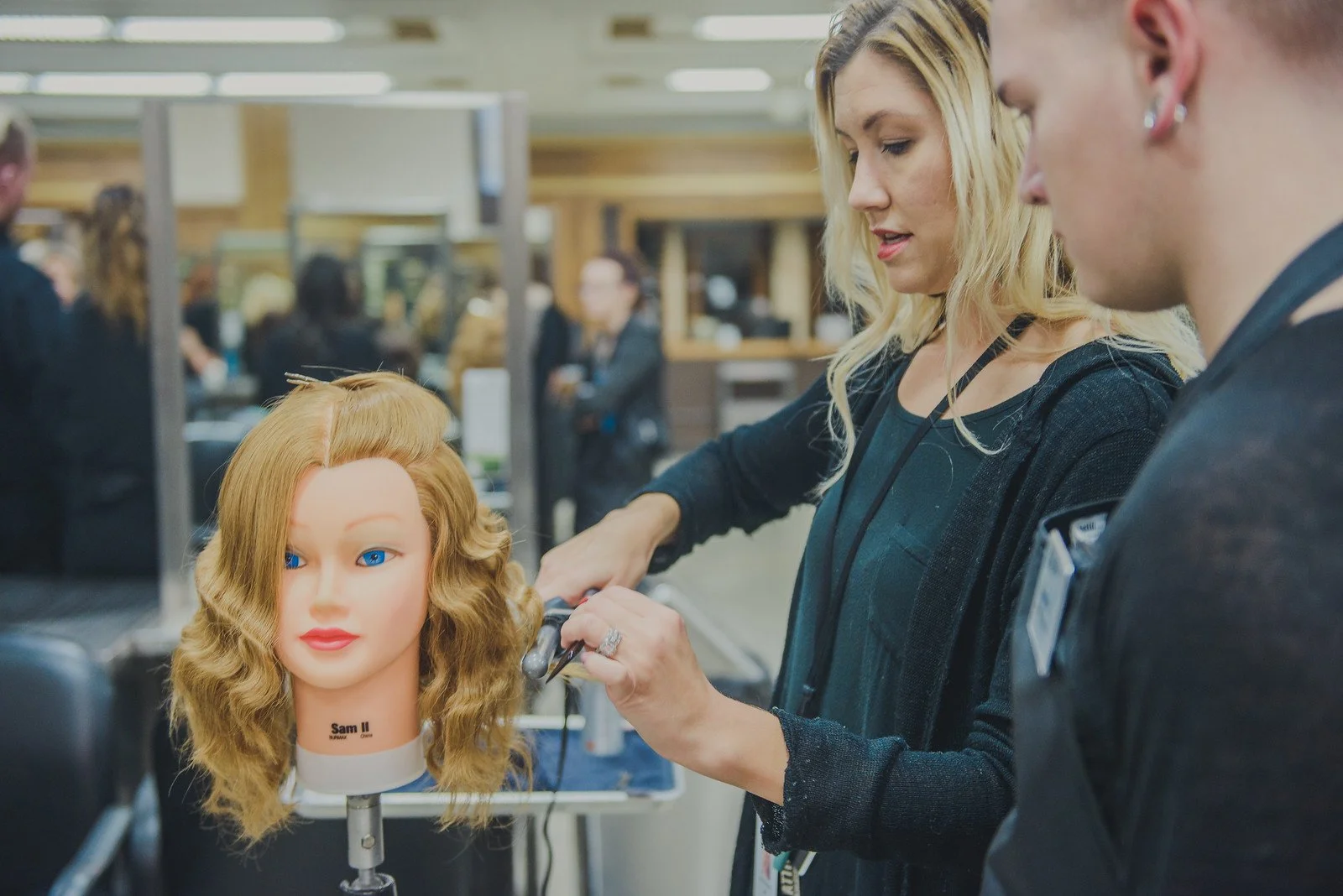 A woman hairstylist practicing styling on a hair mannequin head with blonde, wavy hair in a busy hair salon.