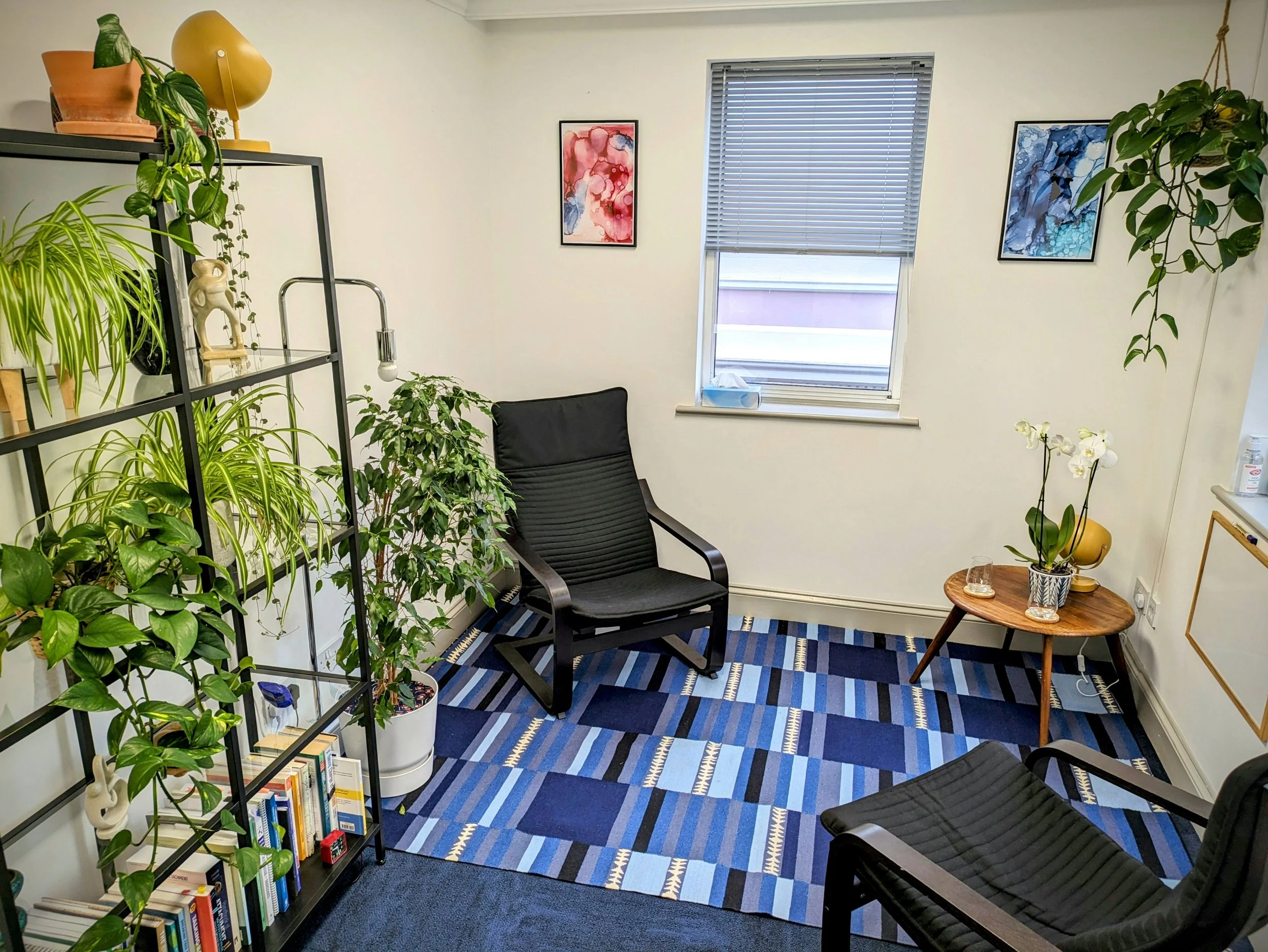 A small office or lounge with a black armchair, a black chair, a small wooden side table with orchids, a bookshelf with books and plants, and several framed abstract artworks on the walls. The room has a window with closed blinds and a blue geometric patterned rug.