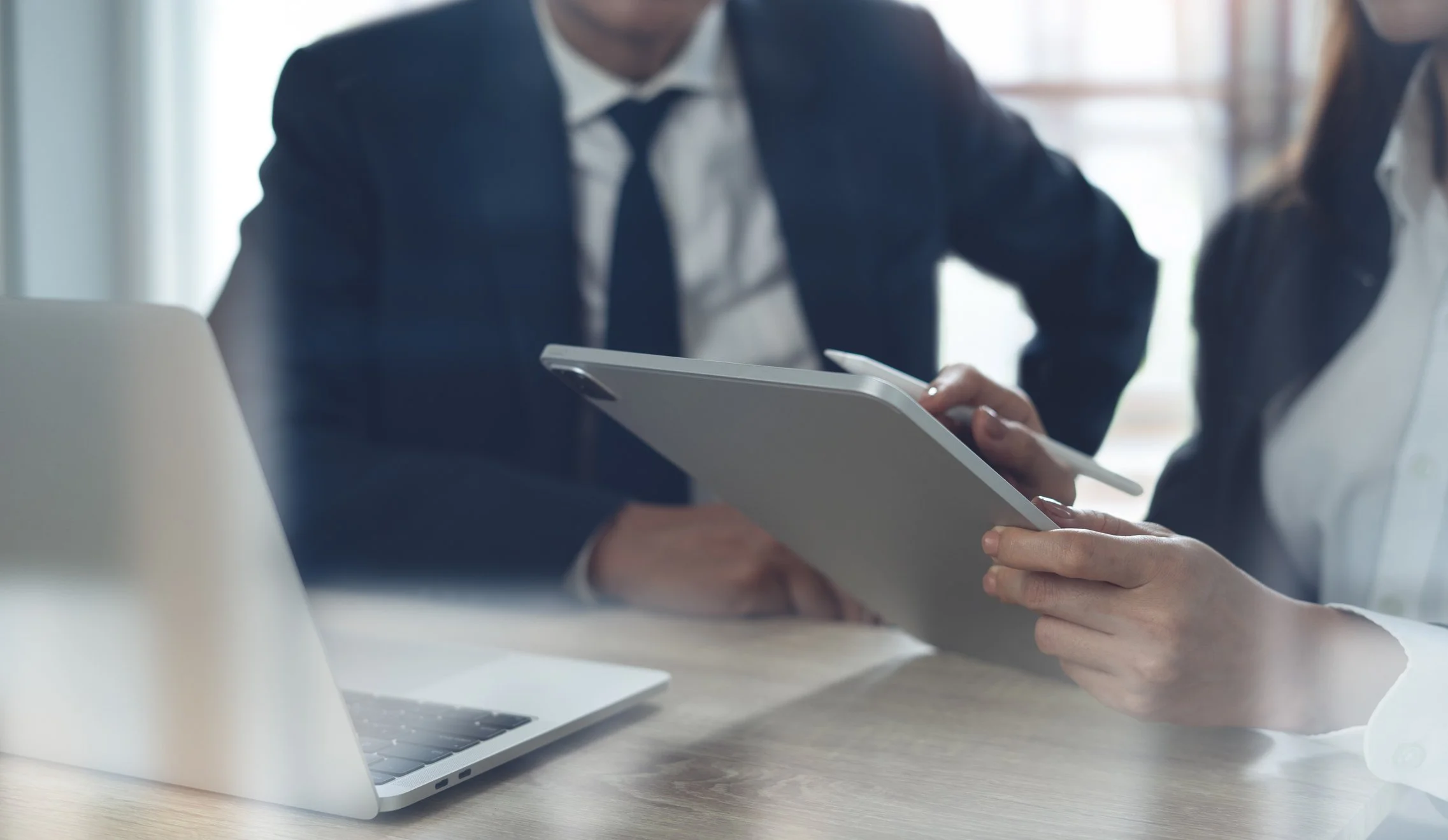 Zwei Geschäftsleute in Anzügen arbeiten an einem Tablet, mit einem Laptop auf dem Tisch.