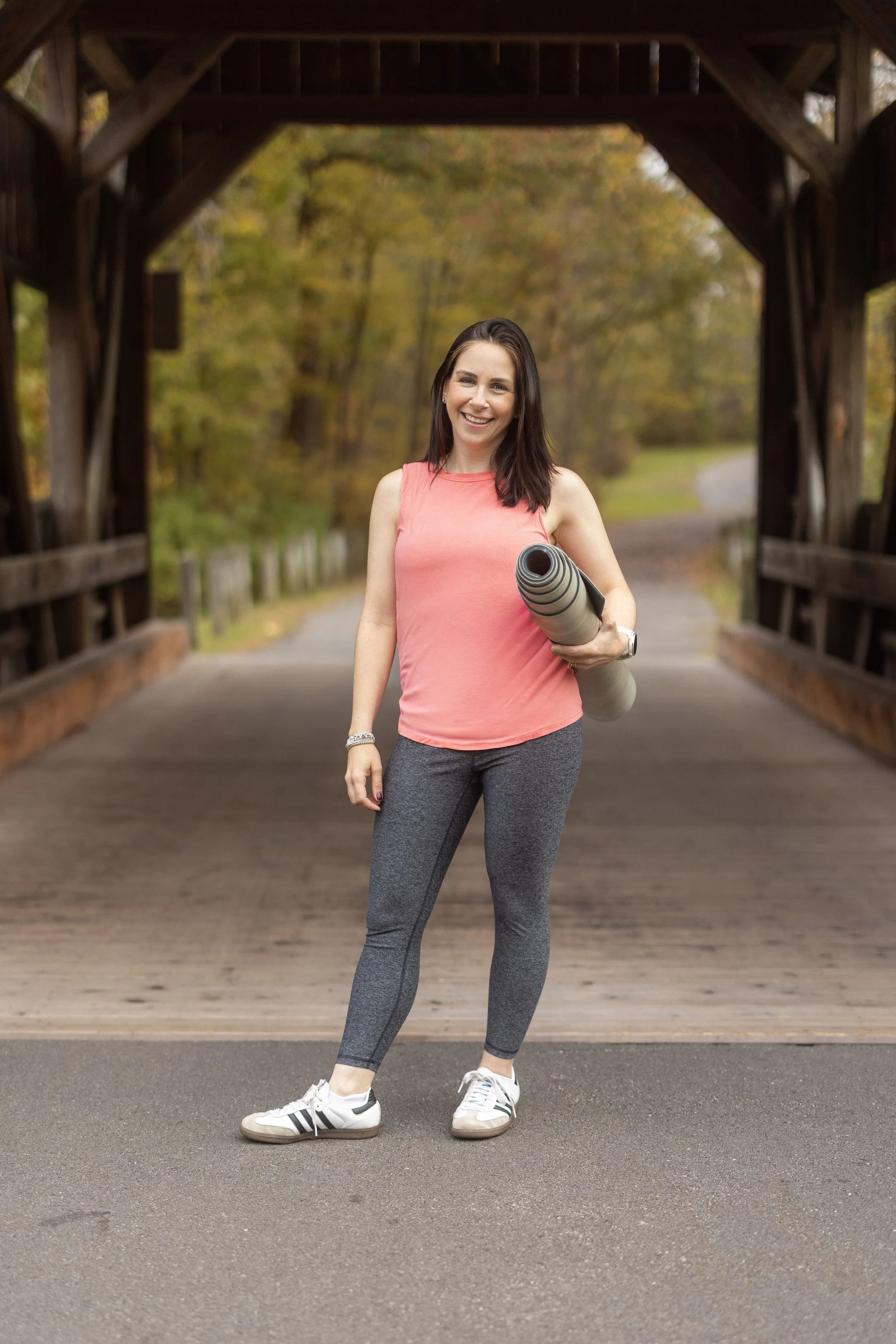 A woman standing on a trail under a wooden bridge, holding a rolled-up yoga mat, smiling outdoors in fall.