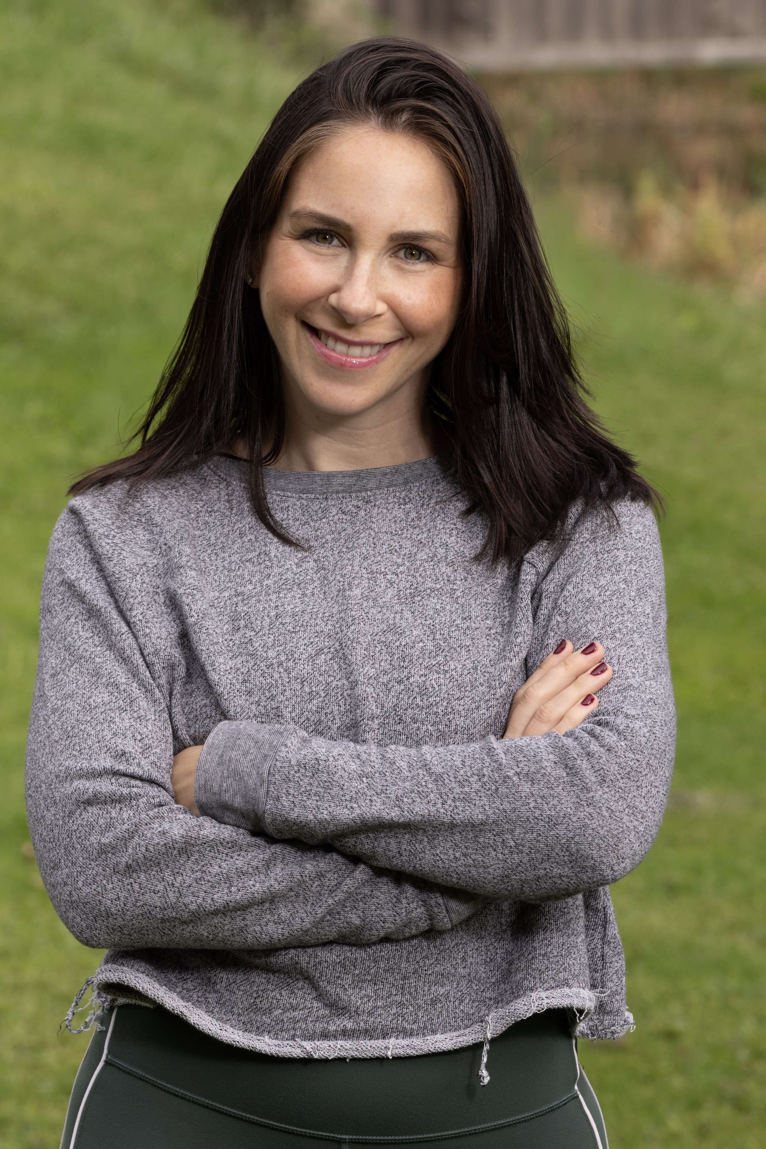 A woman with dark brown hair smiling outdoors, wearing a gray sweater and black pants, standing on grass with a blurred background of greenery.
