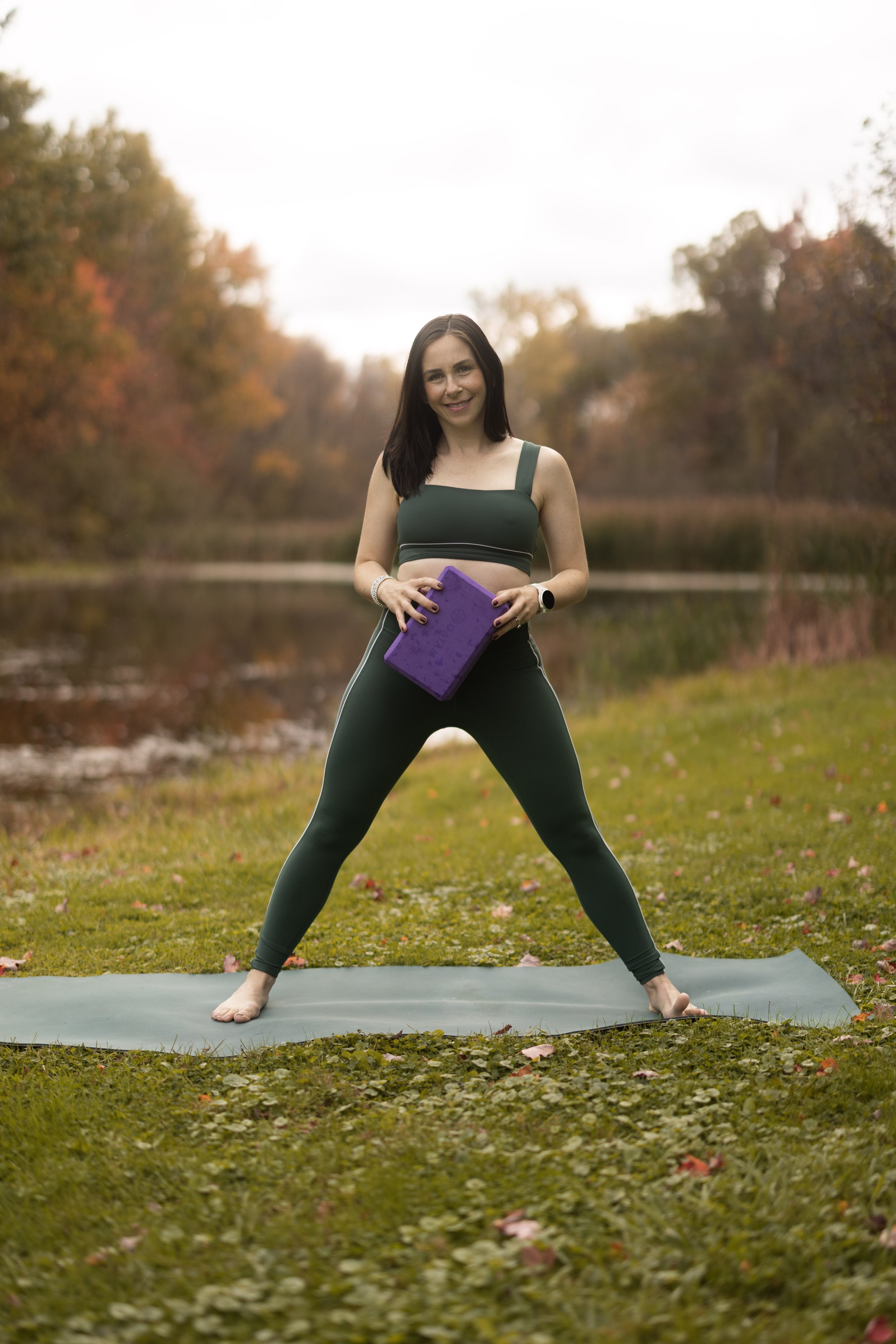 Woman practicing yoga outdoors in a wooded area with fall foliage, standing on a yoga mat, holding a purple yoga block, wearing a green sports bra and leggings, smiling at the camera.