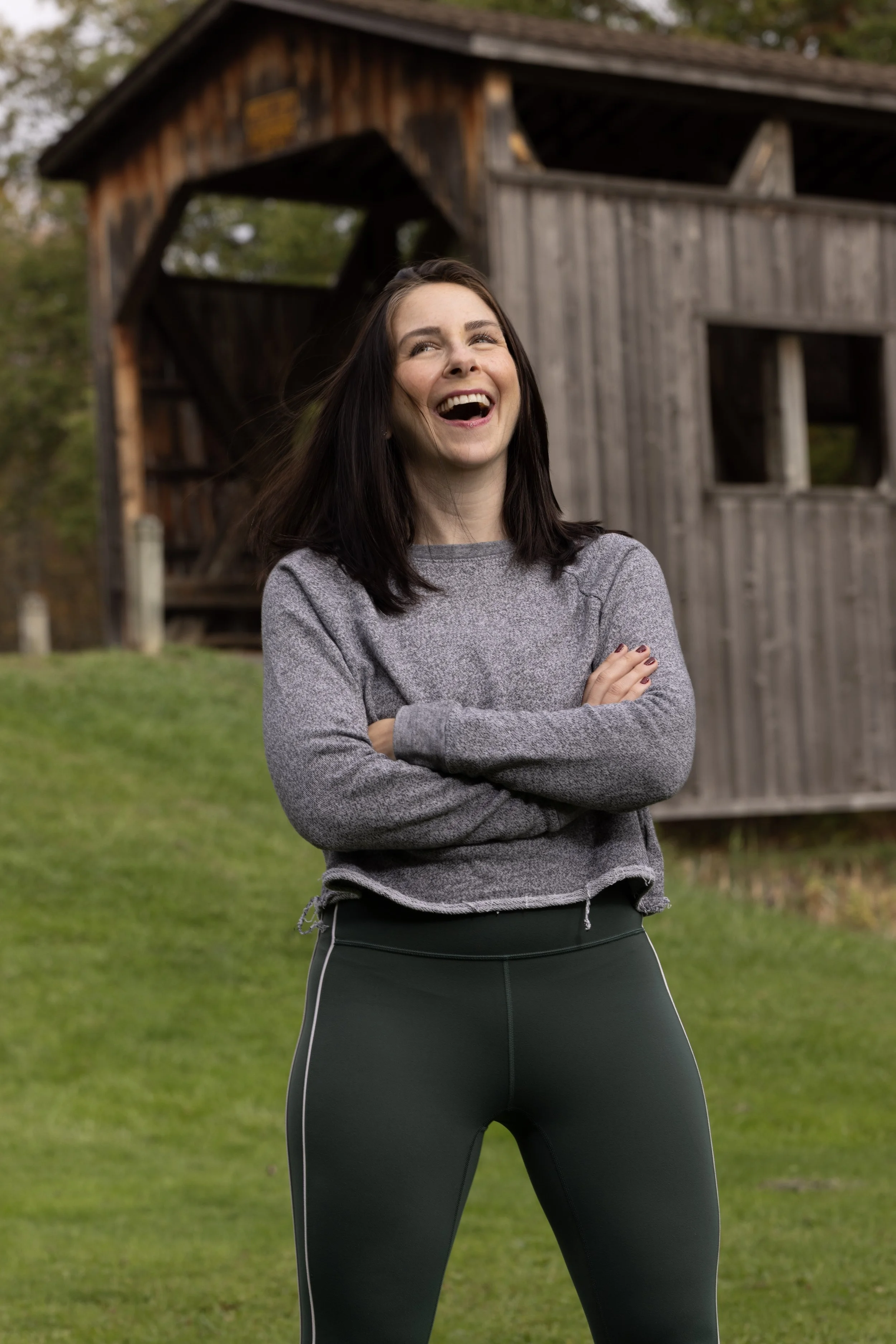 A woman with dark hair outdoors, smiling and standing with her arms crossed, in front of a wooden barn structure and green grassy area.
