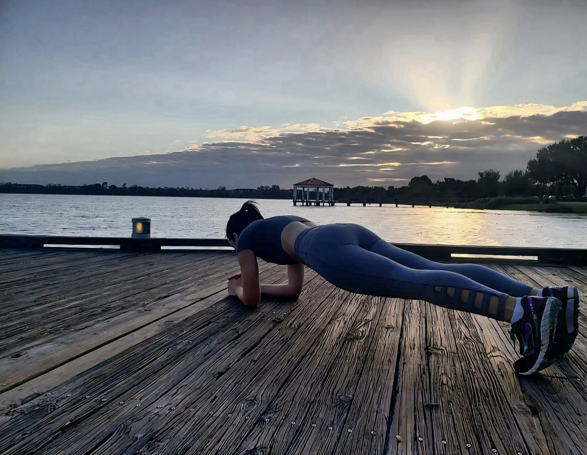 Woman in athletic clothing performing plank exercise on wooden dock by lake at sunset.