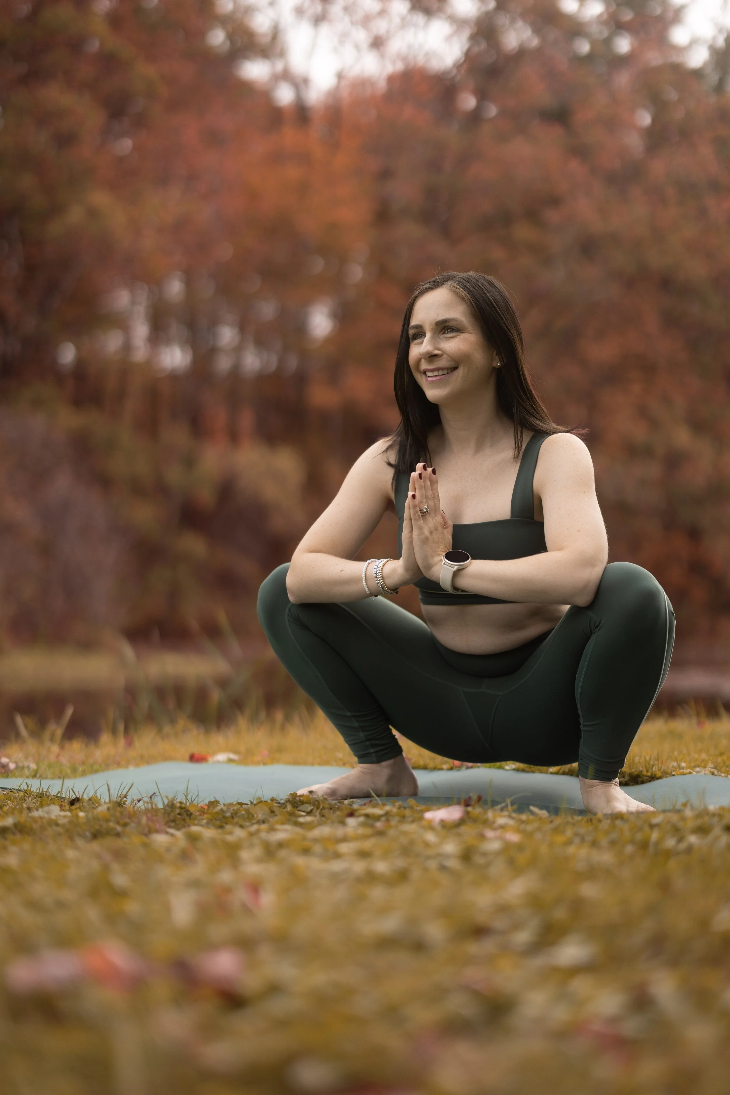 Woman practicing yoga outdoors in autumn, squatting on a yoga mat with hands in a prayer position, smiling, with fall trees in the background.