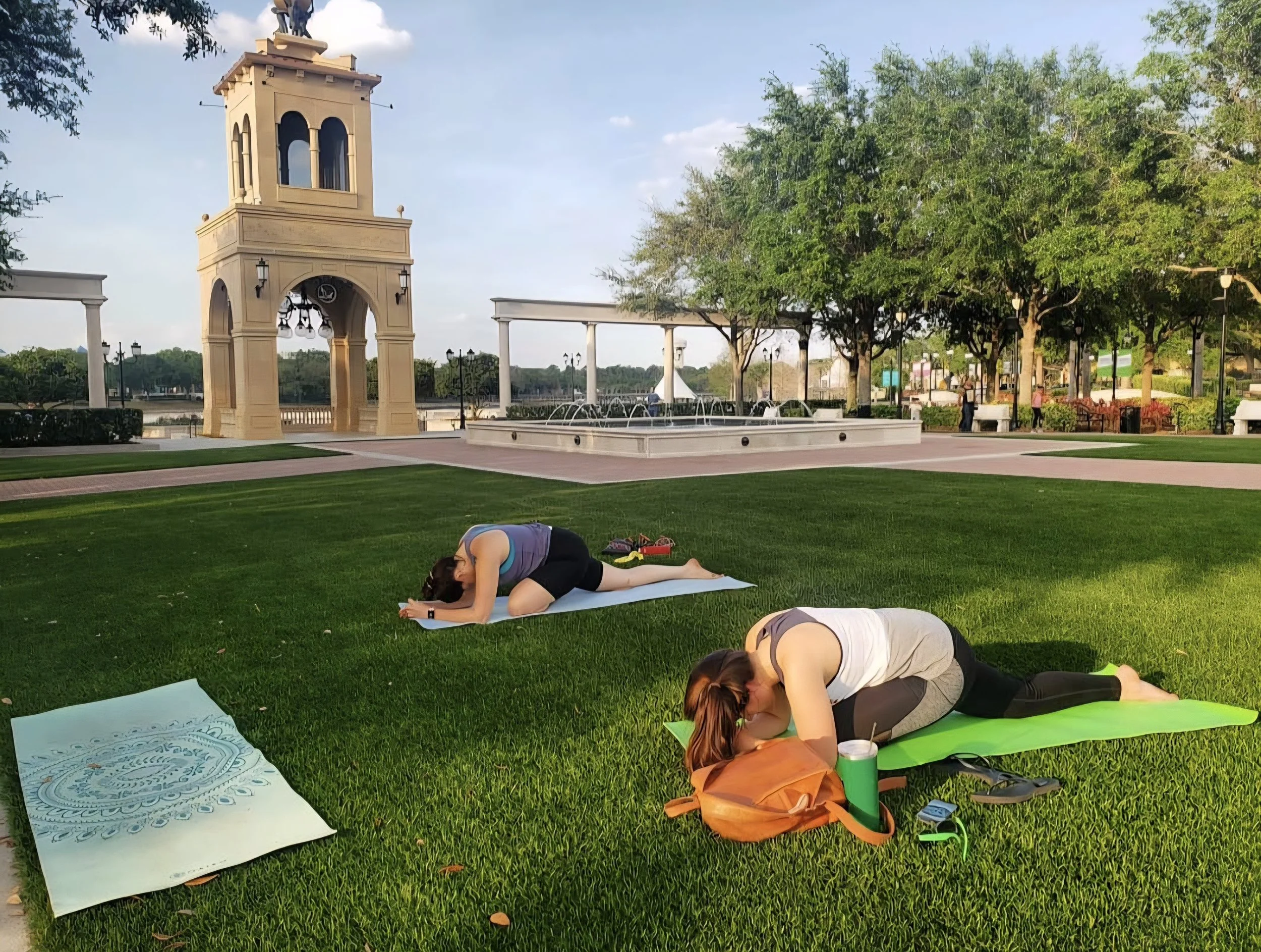 Two women practicing yoga outdoors in a park with a fountain, trees, and a tower in the background. One woman is on a blue yoga mat, the other on a green yoga mat, both in a child's pose position.