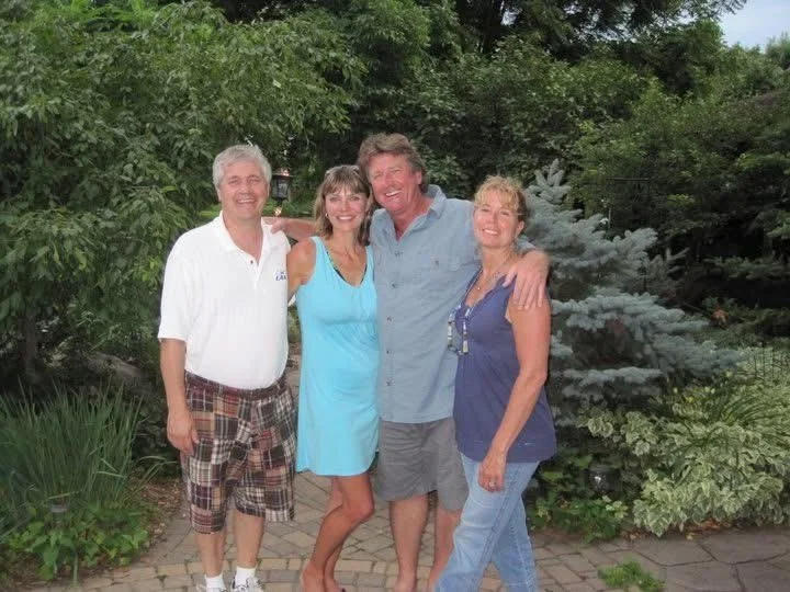 Four people smiling and standing together outdoors in front of greenery, with two men and two women.