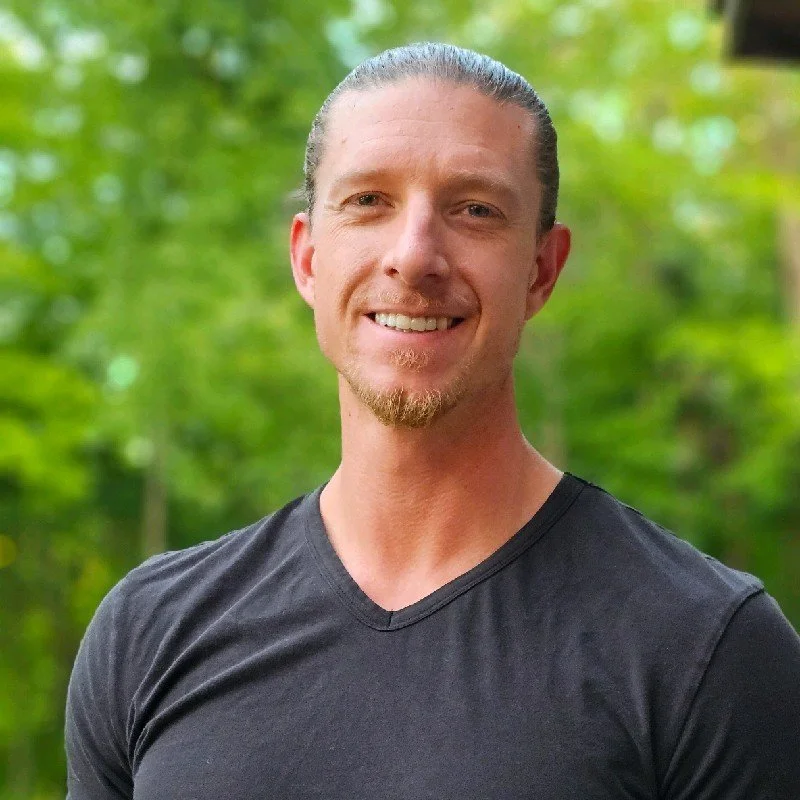 A smiling man with light skin, slicked-back blonde hair, and a goatee, wearing a black V-neck T-shirt, outdoors with green trees in the background.