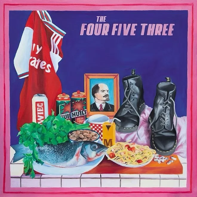 Collection of merchandise and food items on a surface, with a blue wall and a pink frame in the background. Items include a red shirt, cans of beer, a framed portrait of a man, a pair of black boots, a fish with greens, a plate of food, and various other objects. The wall has the phrase "The Four Five Three" written on it.