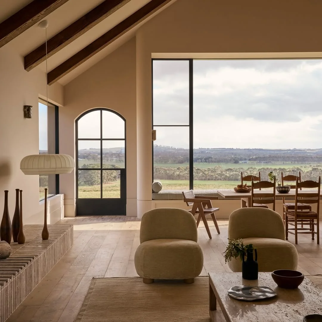 Living room with large windows showing a scenic countryside view, beige chairs, wooden furniture, and minimal decor.