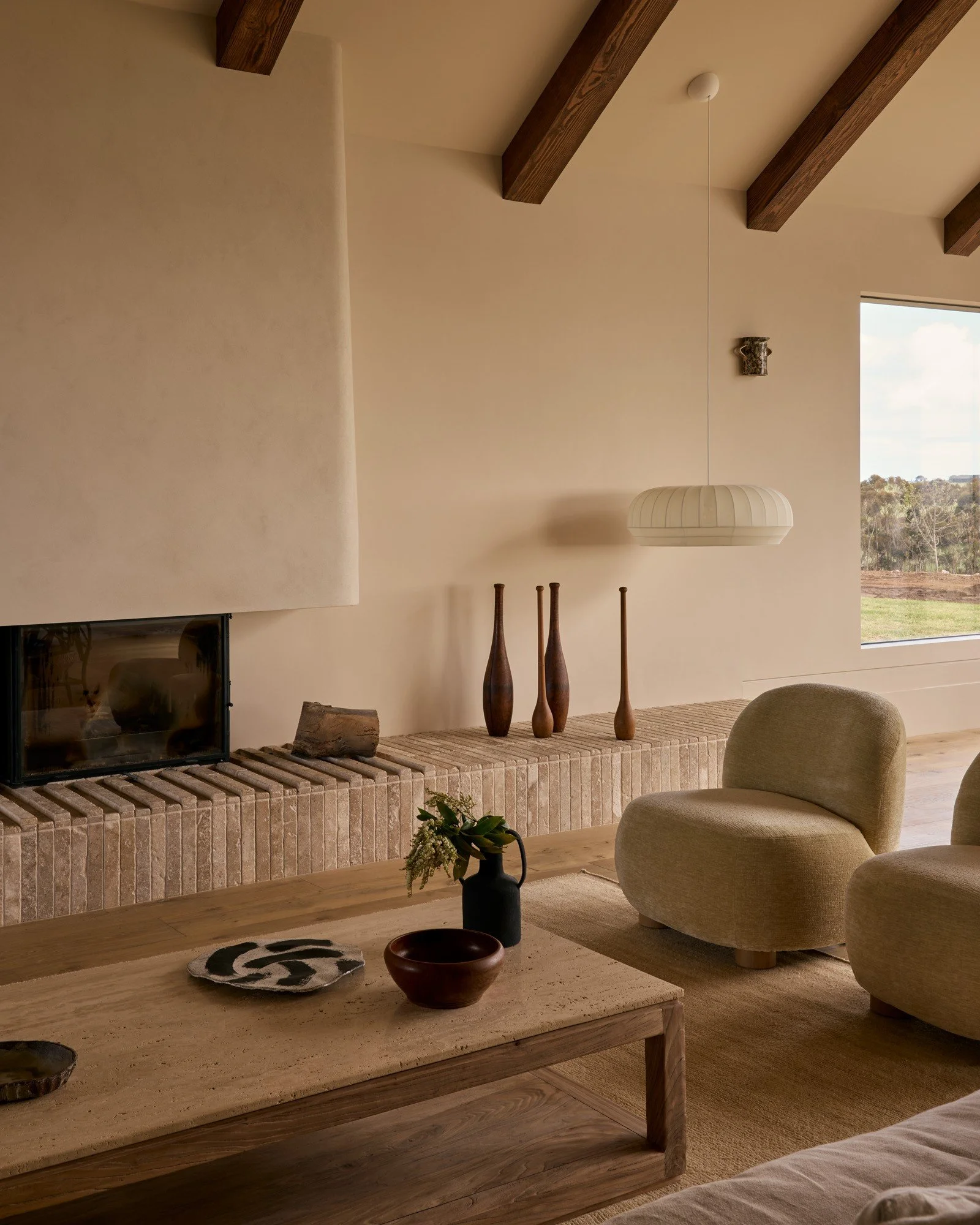A cozy living room with beige walls, wooden ceiling beams, a brick fireplace, beige armchairs, a wooden coffee table with decorative objects, and a window showing an outdoor landscape.