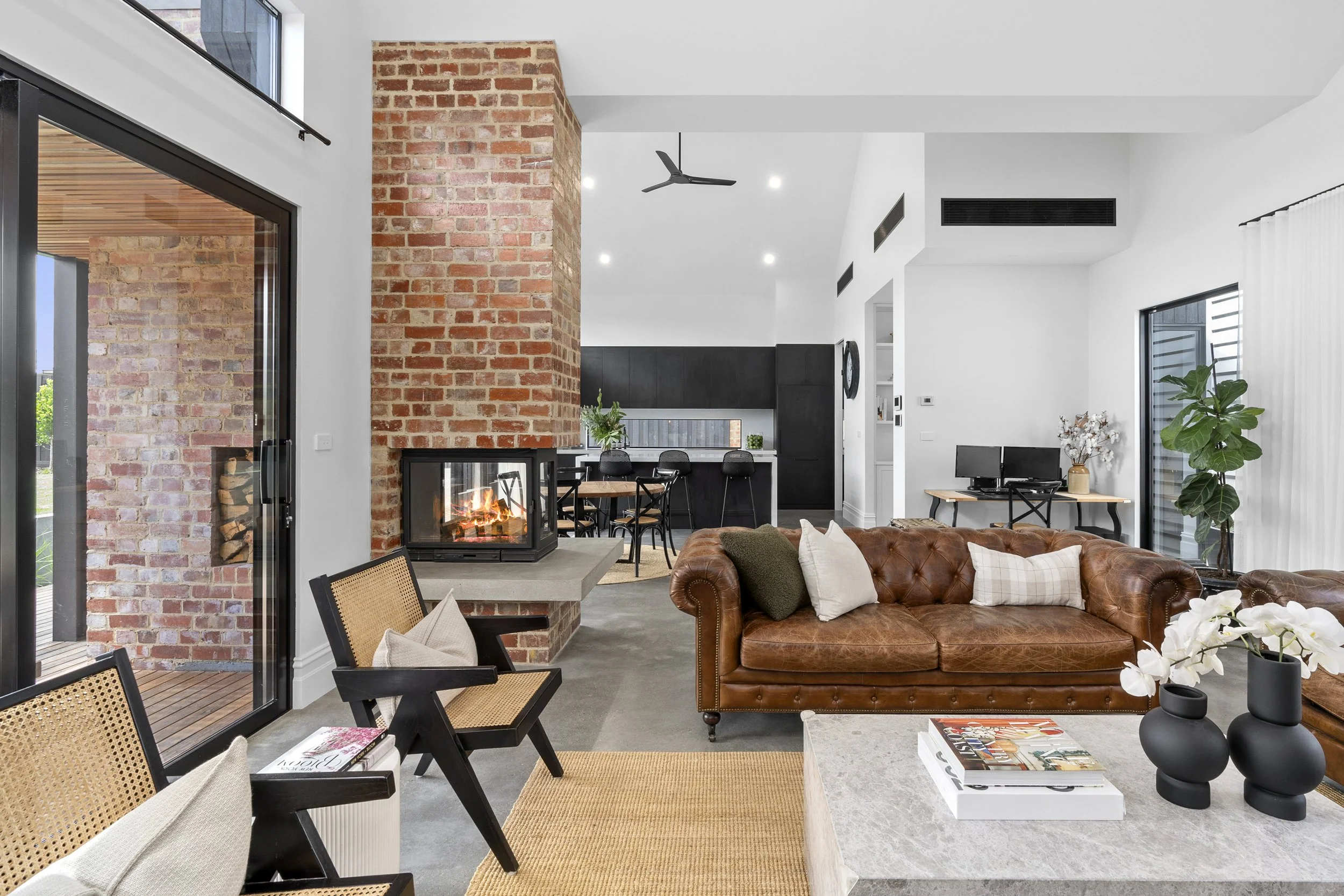 Open-plan living room and kitchen built by Homes By NH featuring brick fireplace, modern black cabinetry, large windows, and leather furnishings.
