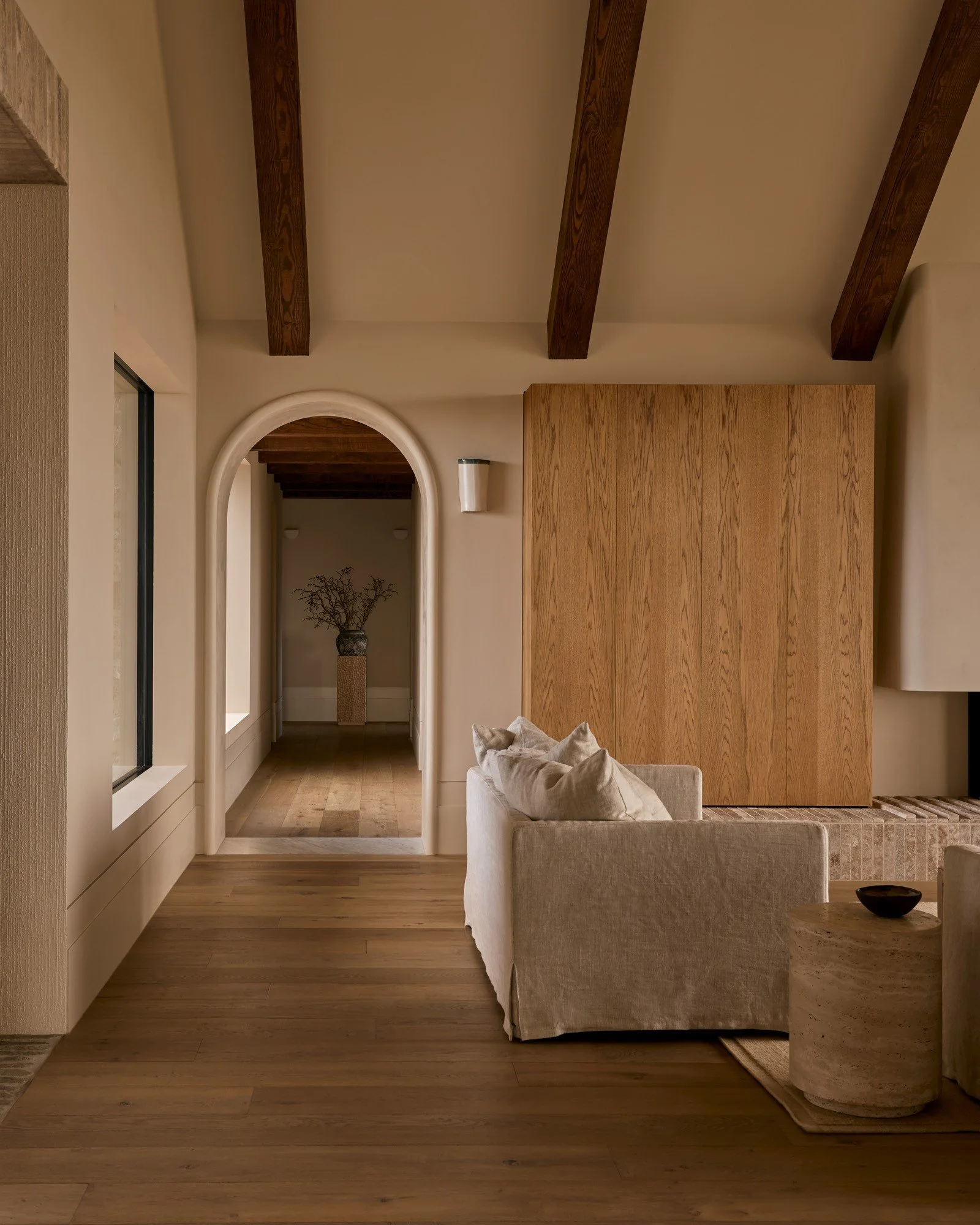Living room with beige sofa, wooden ceiling beams, wooden flooring, and archway leading to a hallway with a decorative branch in a tall vase.