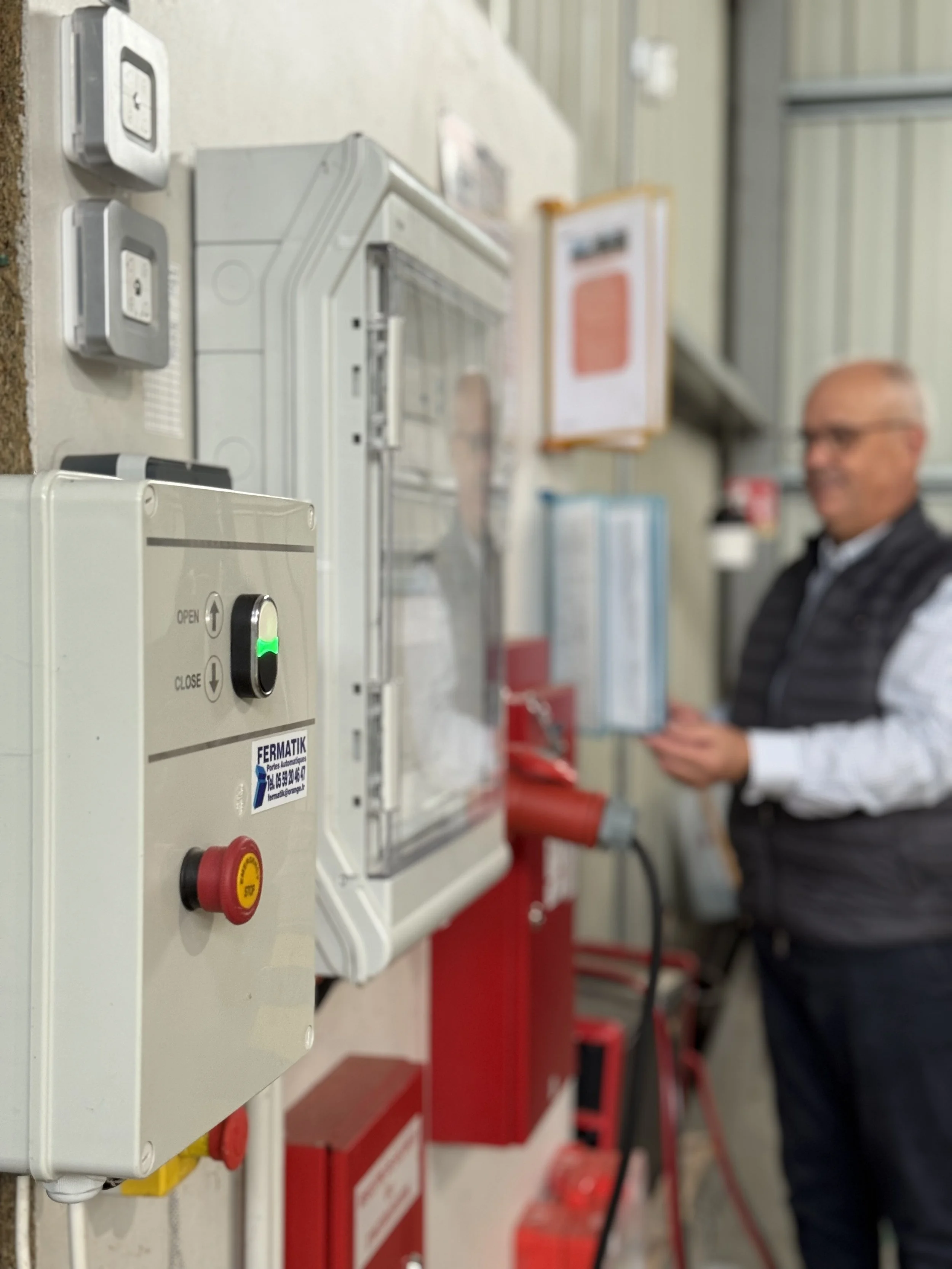 Industrial fire safety equipment mounted on a wall, including a control panel with an emergency stop button, fire alarm, and electrical switches. A man in work attire is seen in the background.