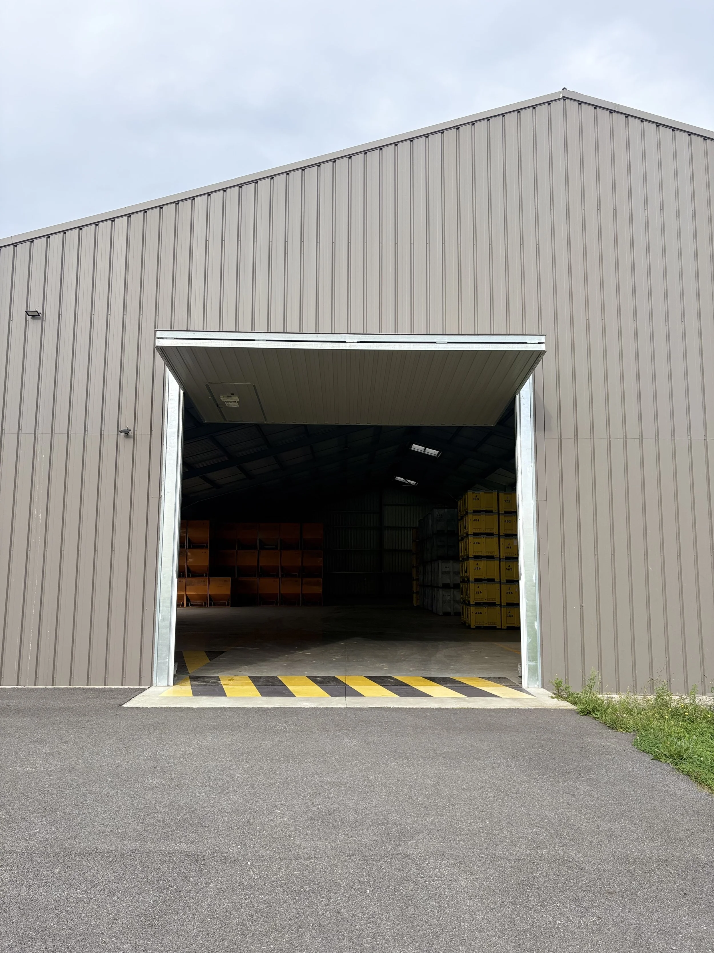 Large industrial warehouse with open garage door, inside stacks of crates and shelves, paved ground in front, overcast sky.