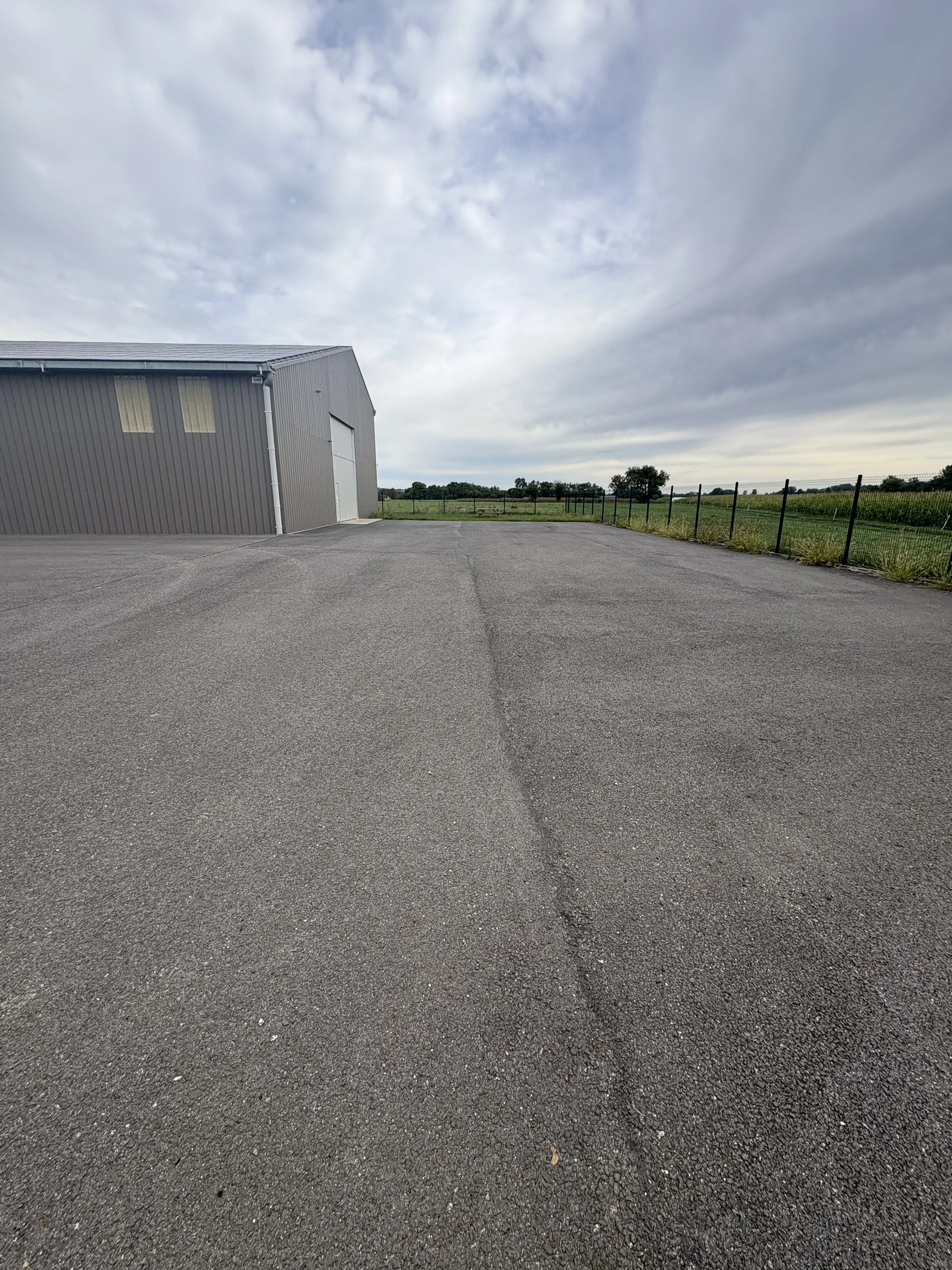 Wide view of an empty paved lot with a large gray industrial building on the left, a chain-link fence on the right, and open green fields with trees in the distance under a mostly cloudy sky.