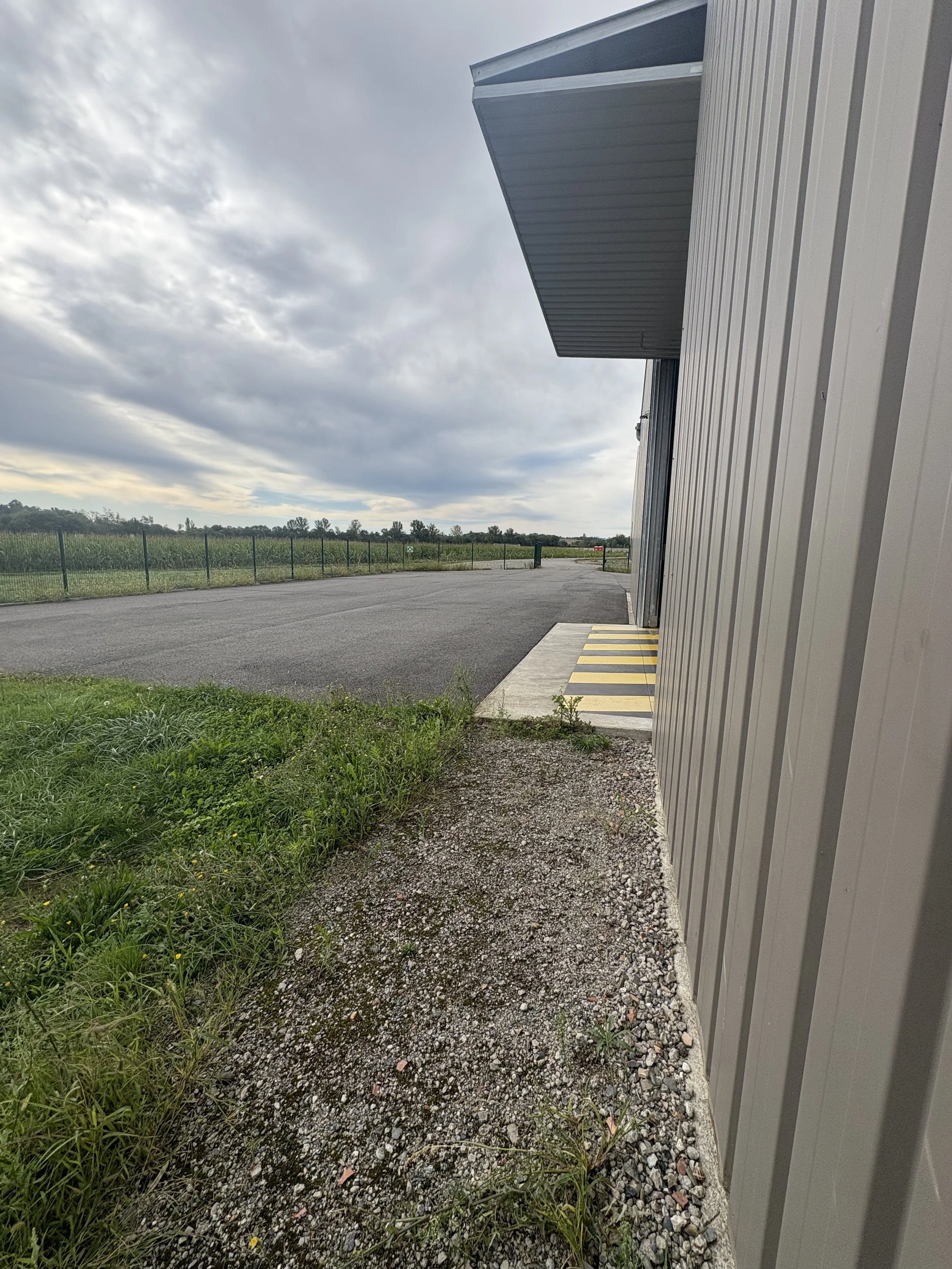 Hotel or industrial building exterior with gravel pathway, green grass, parking lot, and open fields under cloudy sky.
