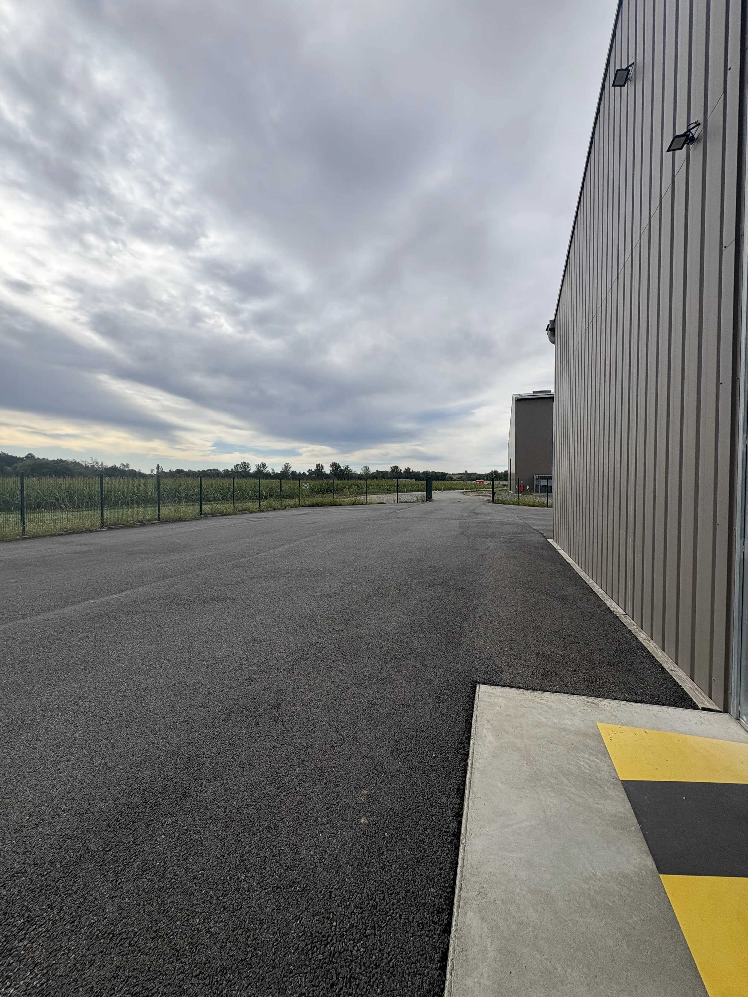 Empty industrial or warehouse parking lot with a large gray building on the right, overcast sky, and open field in the distance.