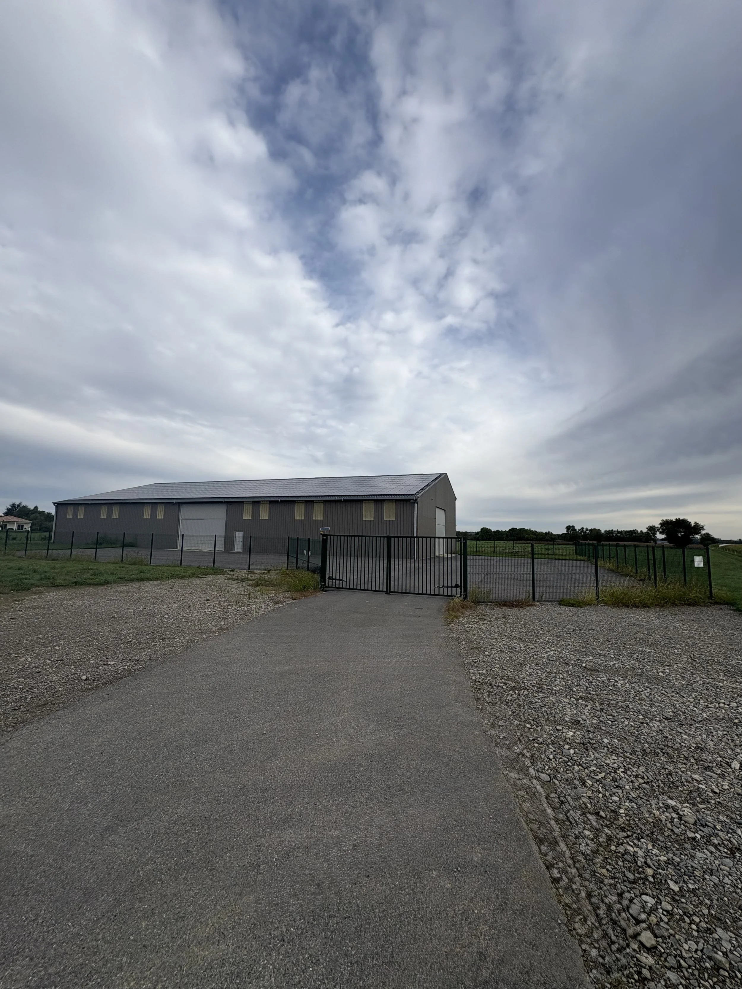 A large metal building with a sloped roof, surrounded by a black fence and gate, on a gravel and paved area under a cloudy sky.