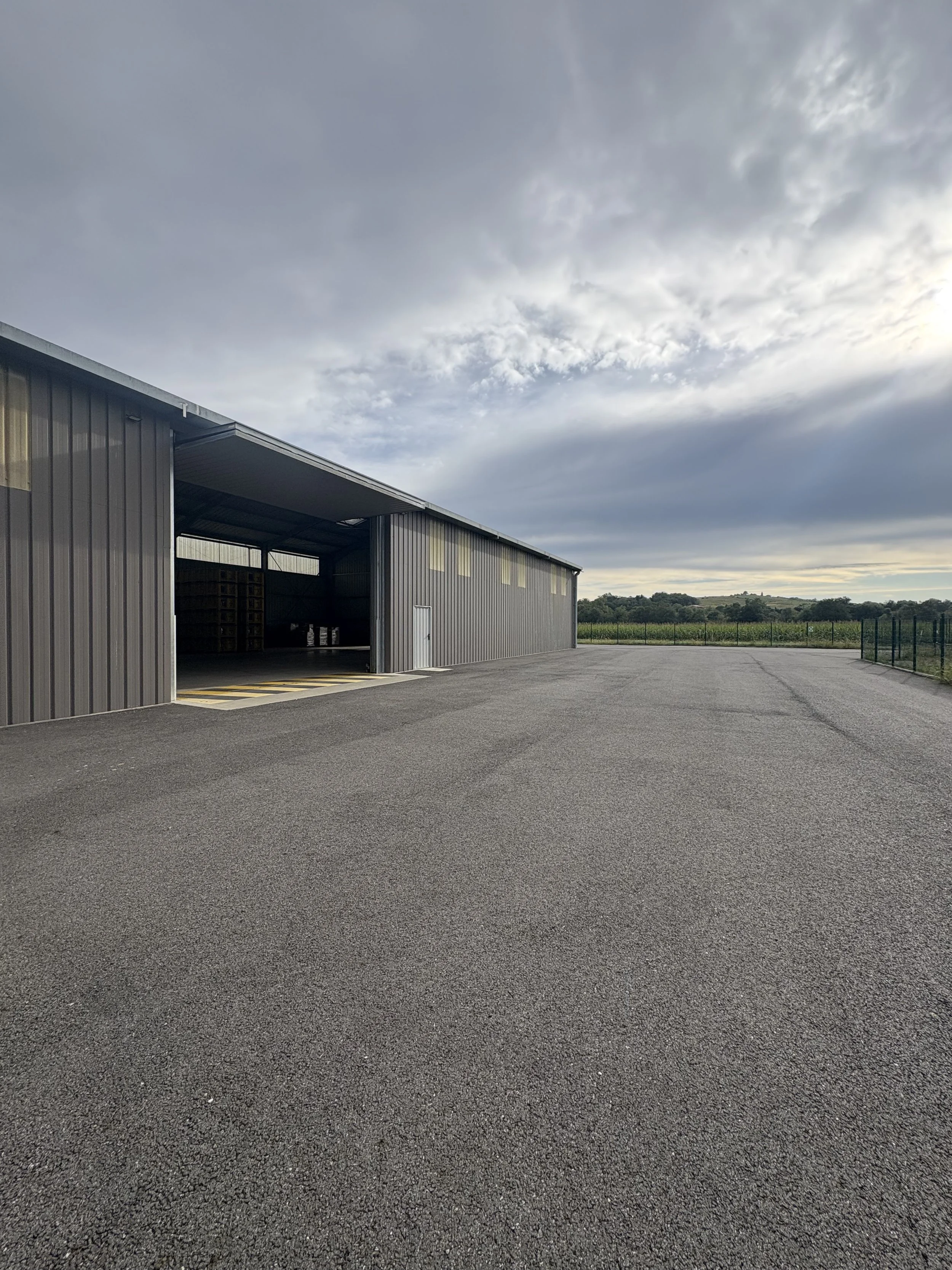 An empty asphalt lot next to a large grey metal industrial building with an open garage door and small windowed sections on the upper part. The sky is partly cloudy with a distant green landscape.