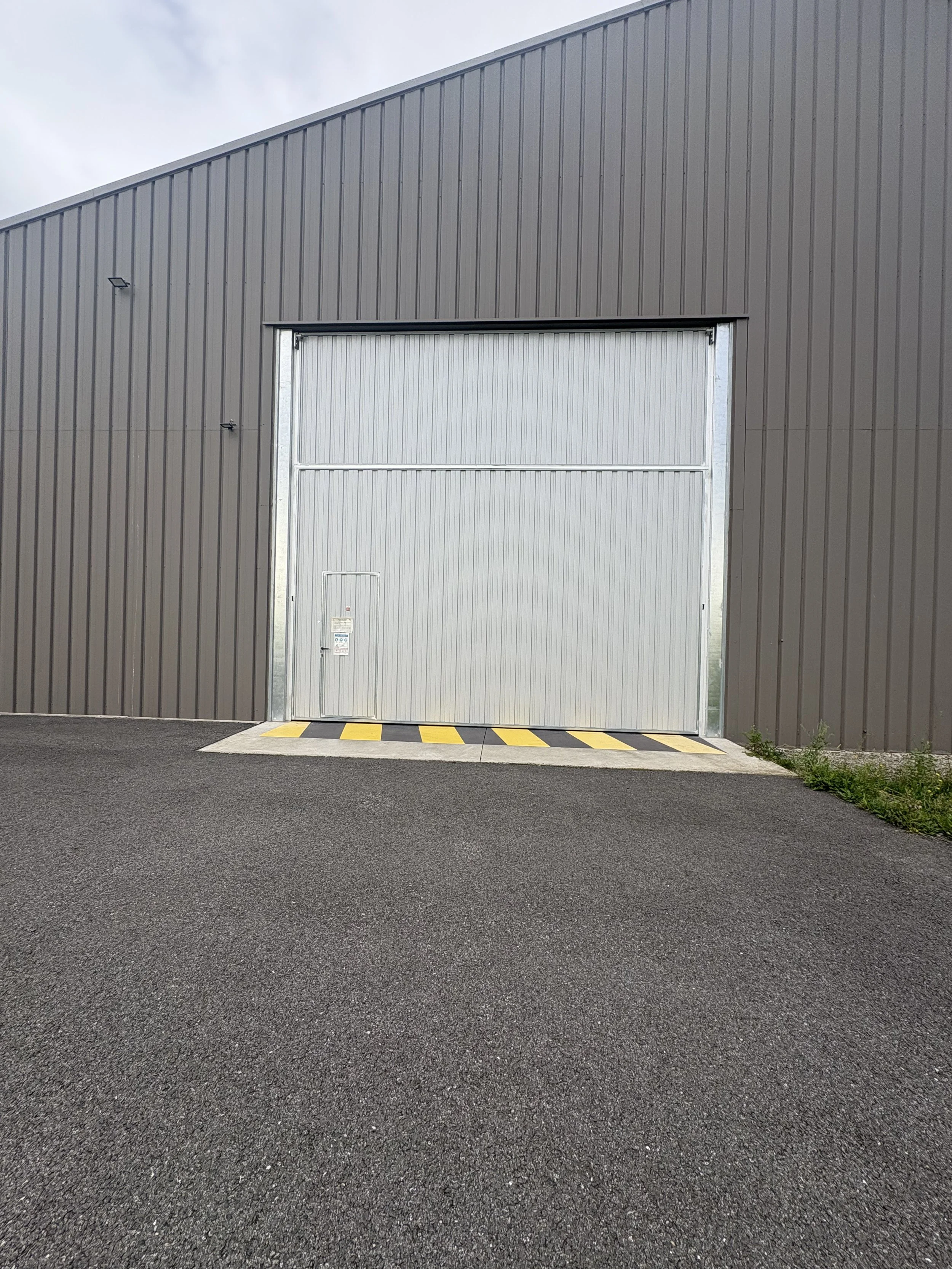 Large gray industrial or warehouse building with a big metal roll-up door, surrounded by a paved asphalt area and some grass at the side.