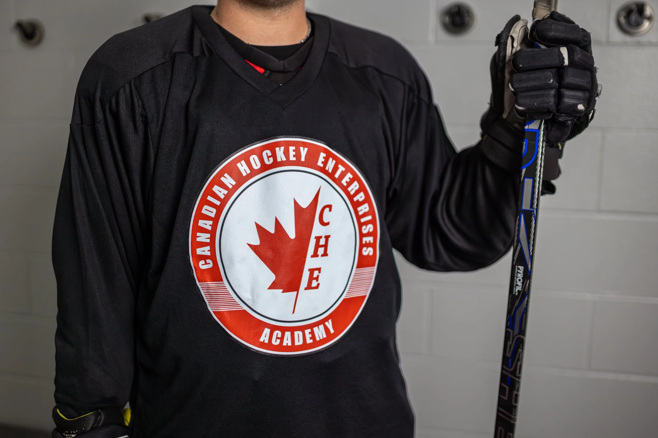 Close-up of a person wearing a black hockey jersey with the Canadian Hockey Enterprises Academy logo, holding a hockey stick, against a background of gray lockers.