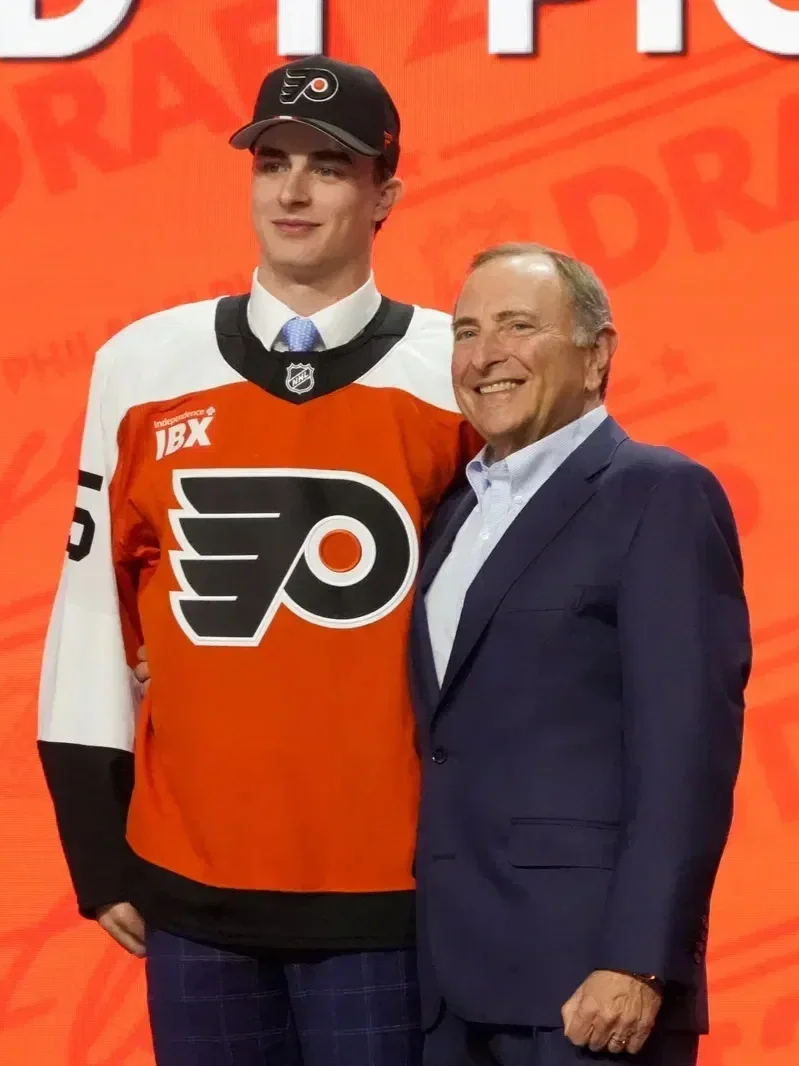 Porter Martone in an Philadelphia Flyers hockey jersey and cap, standing next to an older man in a navy suit and light blue shirt, both smiling in front of an orange background with white and black text.