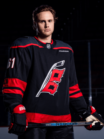 Riley Stillman in a black and red uniform holding a stick with a logo, standing on ice rink.