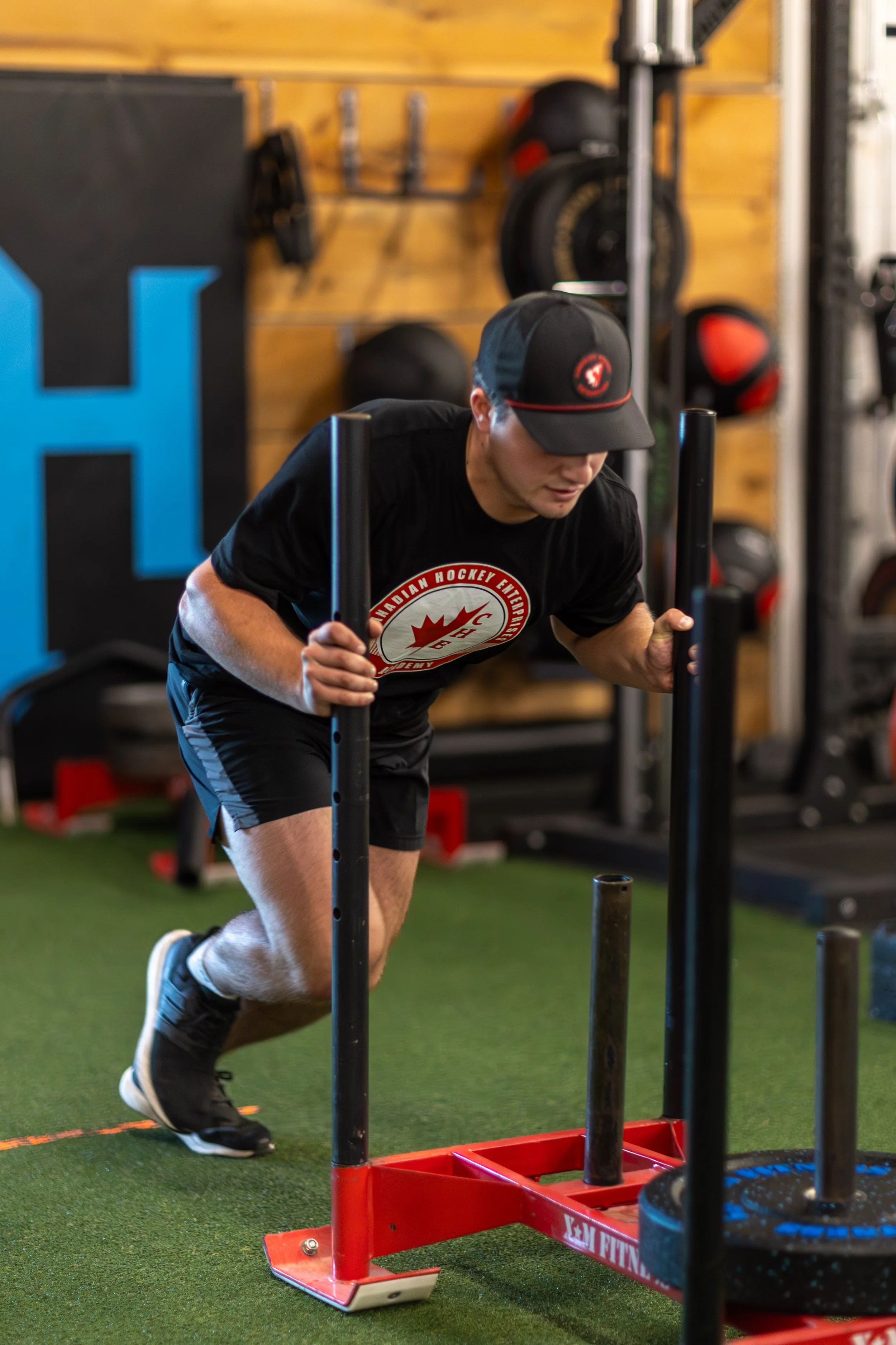 A man wearing a black t-shirt with a logo and a black baseball cap pushes a sled with weights on it in a gym.