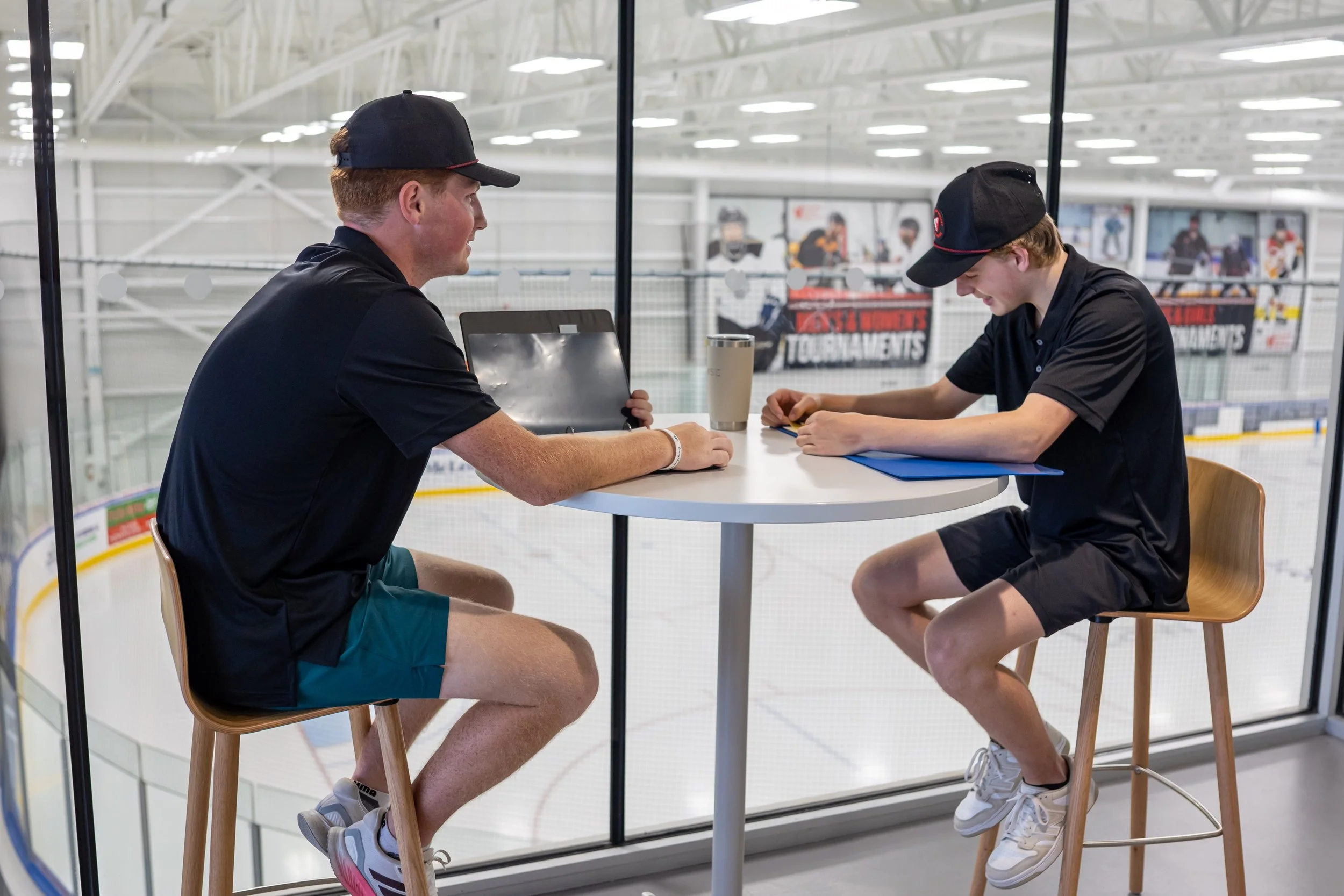 Two young males sitting at a high table with hockey rink visible through glass behind them. One is using a laptop, the other writing or drawing on paper. Both wearing black shirts and caps.