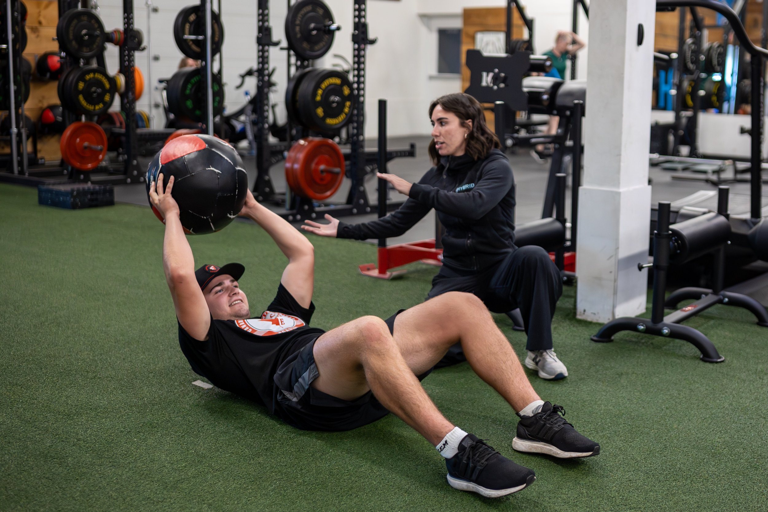 A man lying on the gym floor holding a medicine ball above his chest, with a woman kneeling nearby offering guidance in a gym setting with weightlifting equipment.