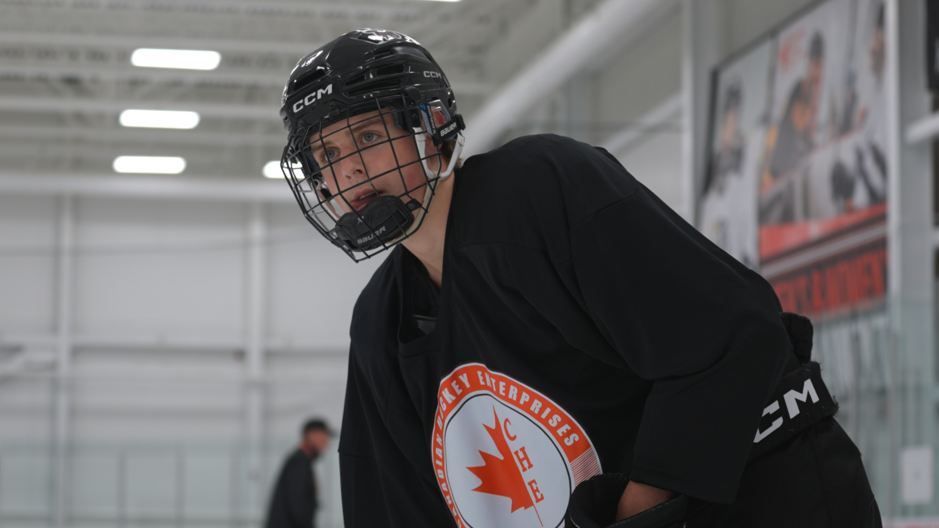 Young male hockey player wearing a black jersey with a logo on the front, black helmet with a face cage, on an indoor ice rink.