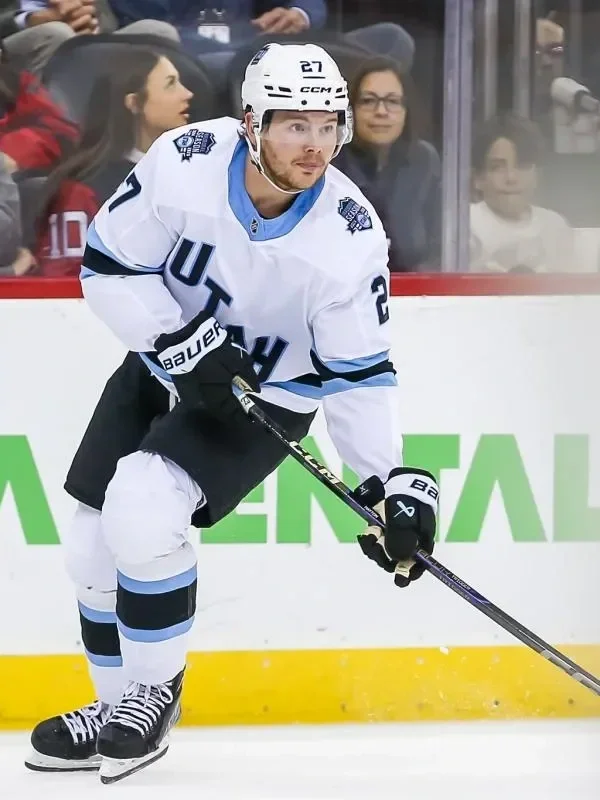 Barrett Hayton in a white jersey with blue and black accents, number 27, bending forward with a hockey stick on the ice during a game. Spectators watch from behind the rink.