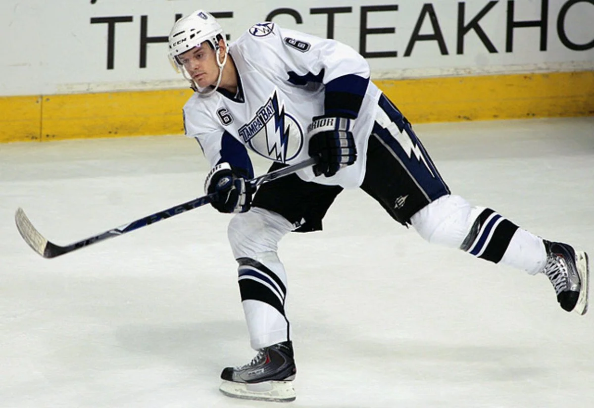 Kurtis Foster in a white and navy uniform skating on the ice, holding a hockey stick, with a yellow barrier and black text in the background.