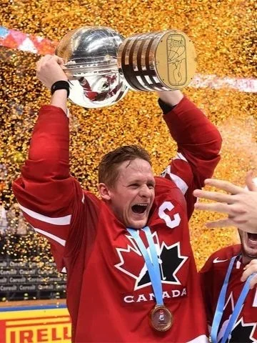 Corey Perry in a red Team Canada jersey celebrating on an ice rink, holding the Stanley Cup trophy overhead, with a gold medal around his neck.