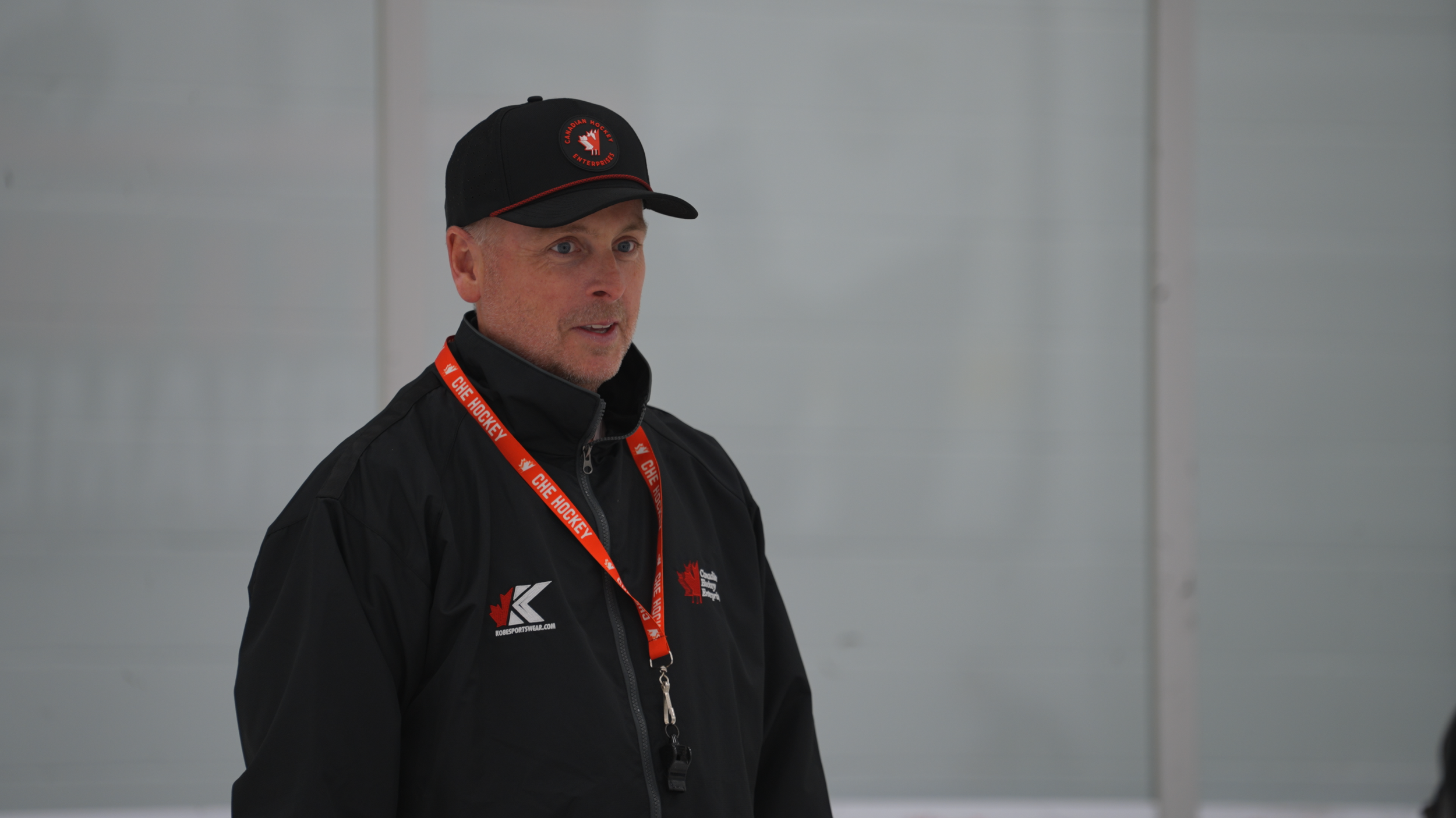 A man wearing a black sports jacket and a black cap with a red maple leaf emblem, standing indoors against a white wall. He has a red lanyard around his neck with white text and is looking to the left.