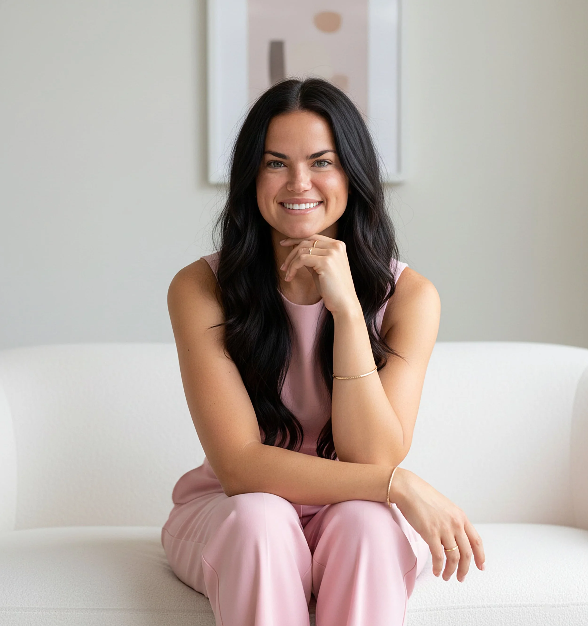 A woman with long dark hair wearing a pink sleeveless top and pink pants, smiling and sitting on a white bench with one hand resting on her chin, in a modern, minimalist room.