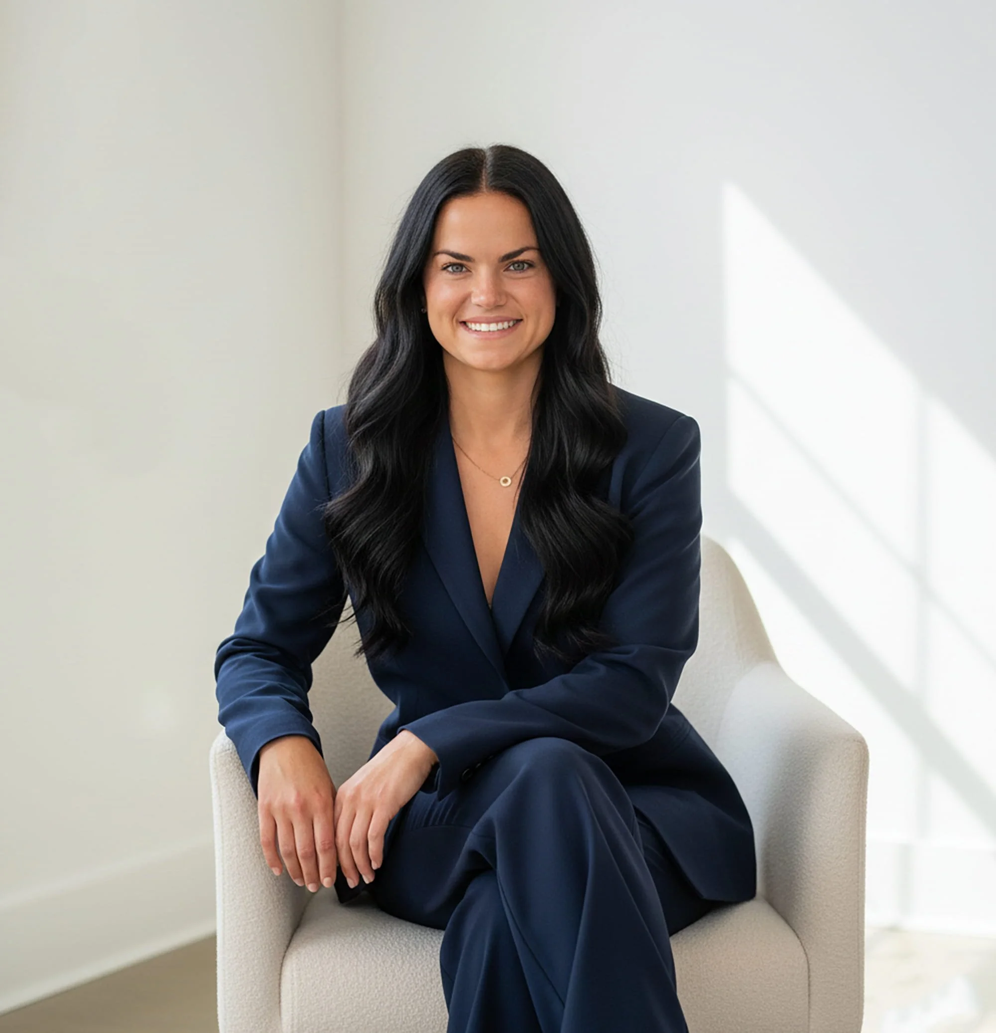 A woman with long dark hair smiling, sitting on a beige chair, wearing a navy blazer and matching pants, in a bright room with white walls and sunlight streaming in.