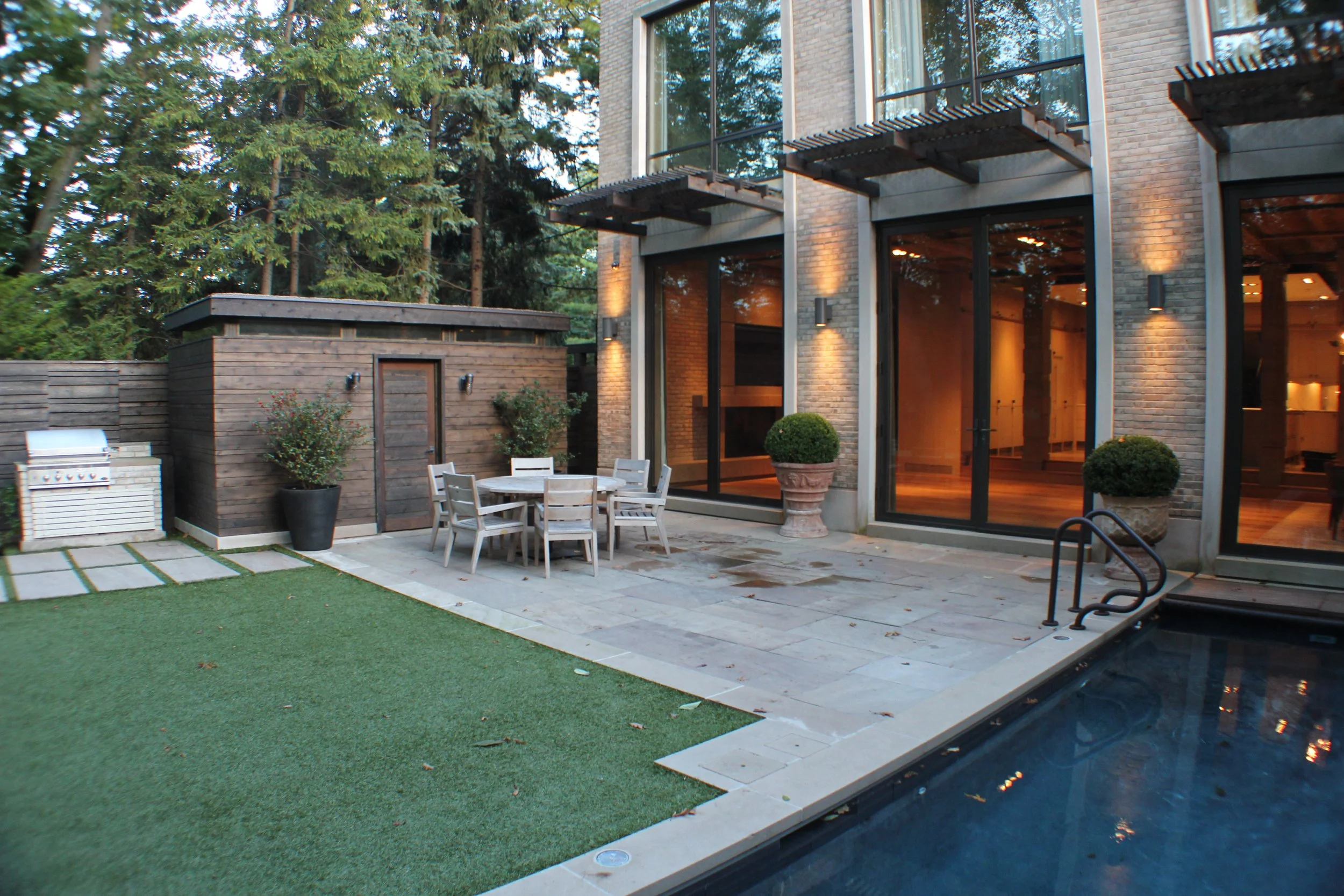 Modern backyard patio with a swimming pool, a round outdoor table with chairs, potted plants, a small wooden shed, and large glass doors leading into a house, surrounded by trees.
