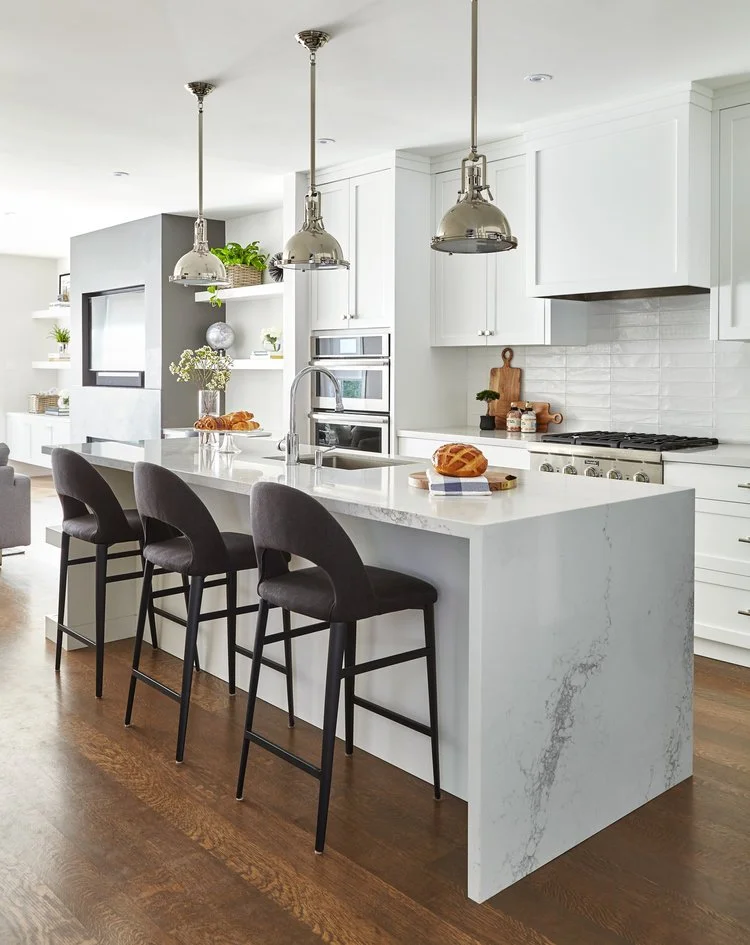 Modern white kitchen with a marble island, three black chairs, stainless steel pendant lights, and wooden flooring.