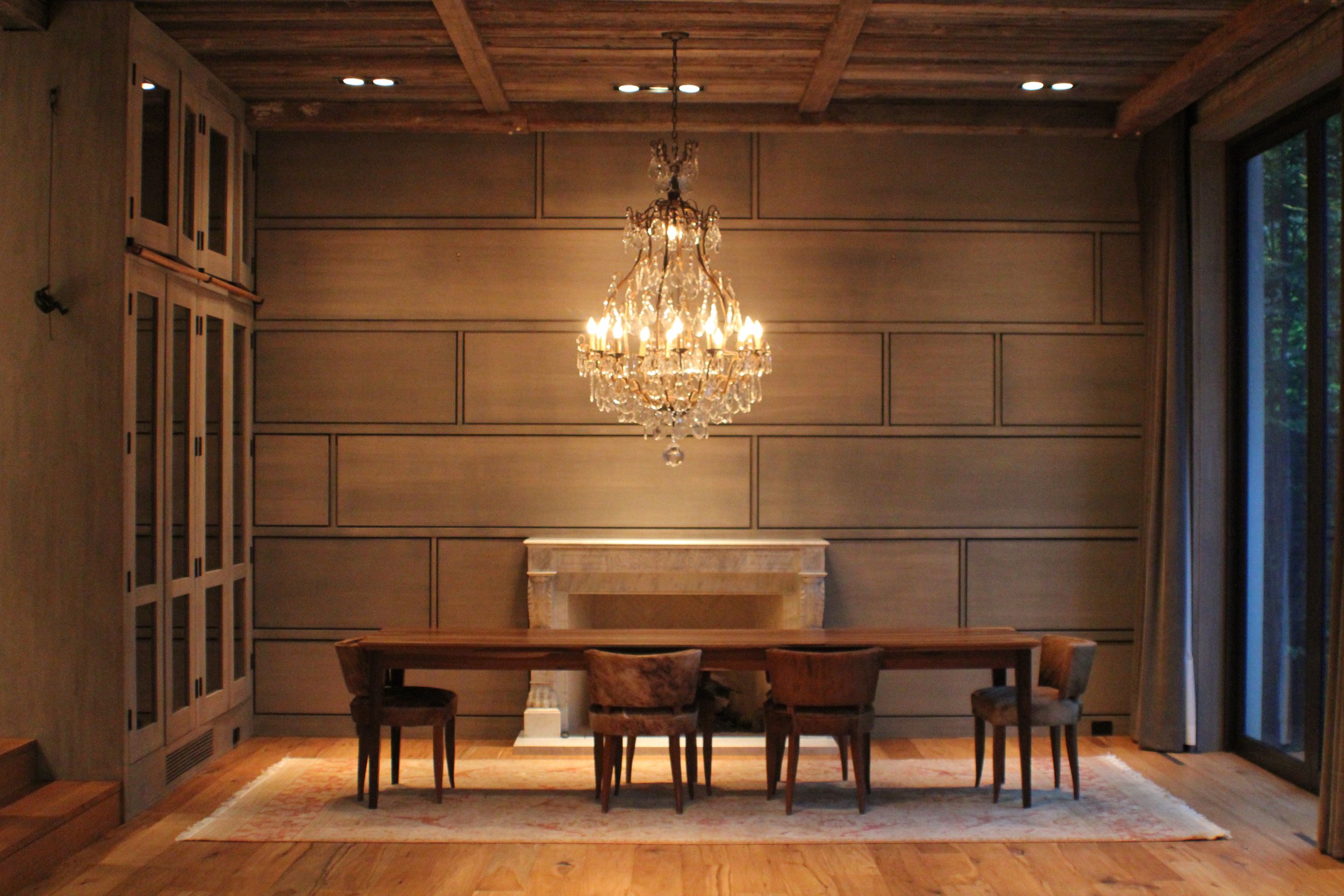 Elegant dining room with a large wooden table, six chairs, a crystal chandelier hanging from a wood-paneled ceiling, a fireplace on the back wall, and large windows with curtains on the right side.