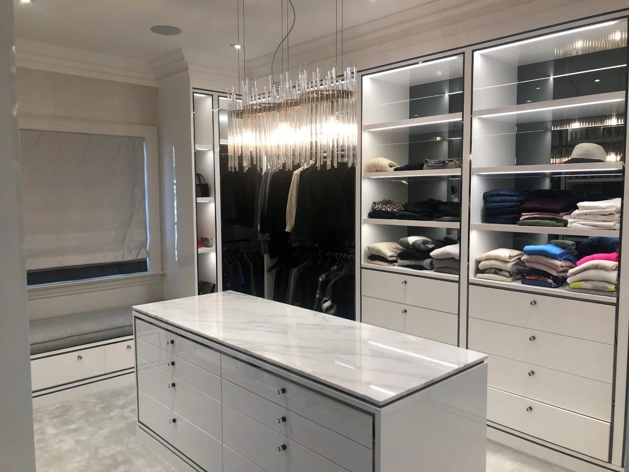 Elegant walk-in closet with white shelving, a marble-topped island, clothing, accessories, and a chandelier.