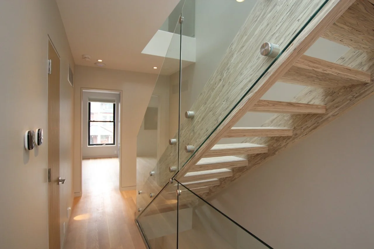 Interior view of a modern stairway with wooden steps and glass railing in a clean, minimalistic apartment hallway.
