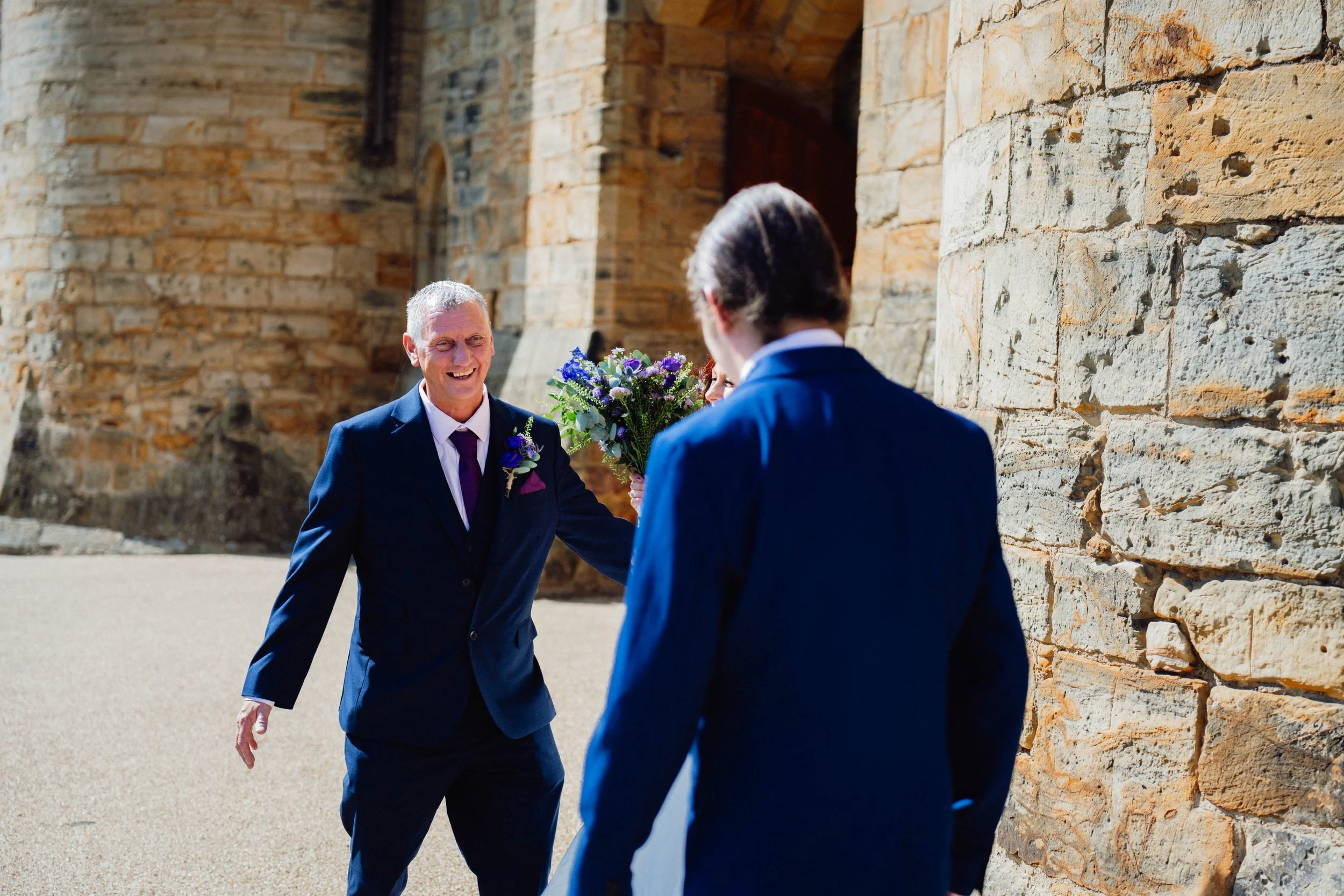 A man in a navy blue suit smiling and holding a bouquet of purple and white flowers, standing outdoors near a stone building, with another man in a similar suit seen from behind.