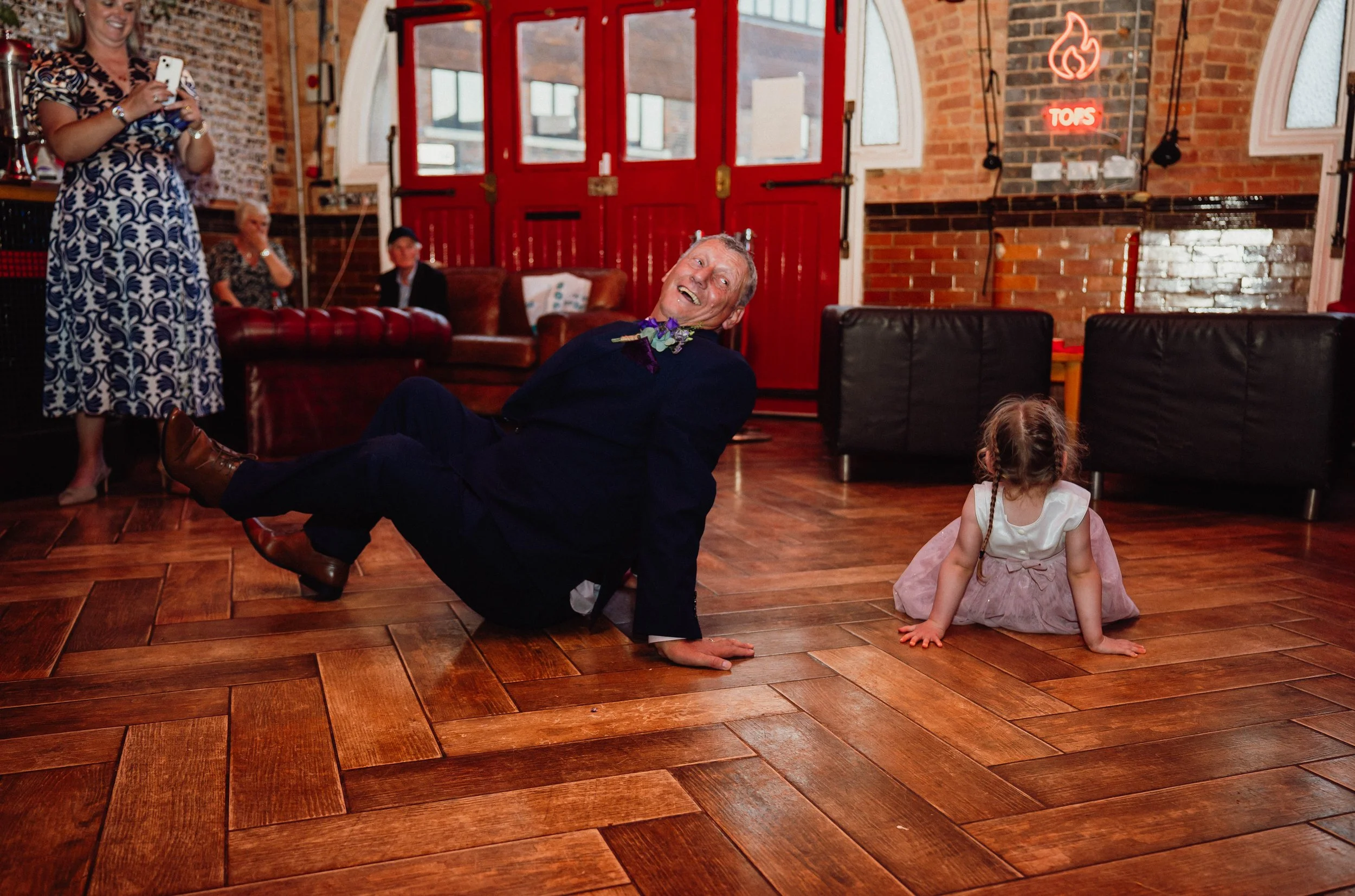 A man in a suit lying on the wooden floor, smiling and looking upwards, with a flower necklace. A little girl in a pink dress sitting nearby with braided hair, on the floor. In the background, a woman in a patterned dress holding a phone, and two wom