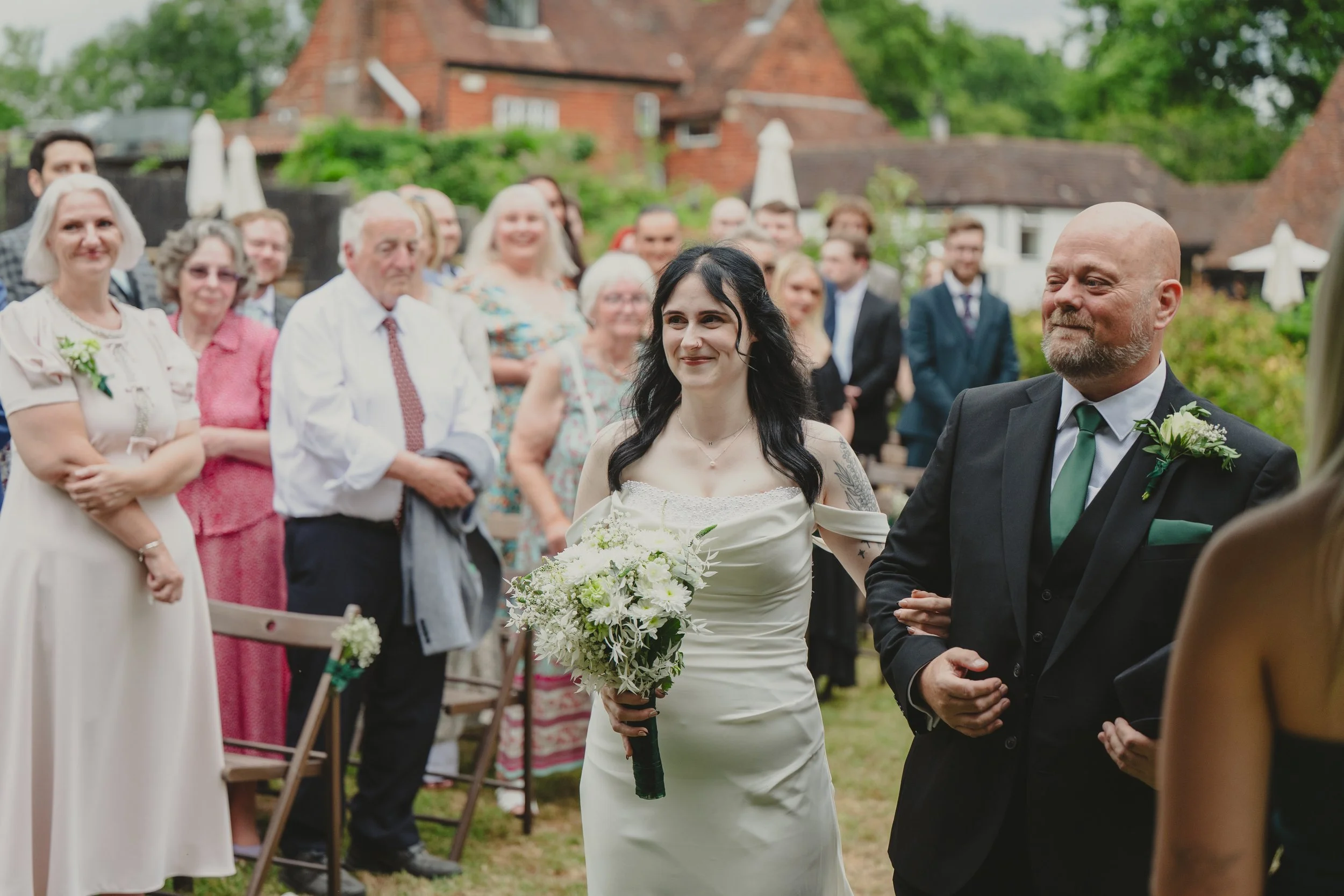 A bride with black hair in a white dress holding a bouquet of white flowers, walking down the aisle beside a groom in a black suit with a green tie, surrounded by family and friends at an outdoor wedding ceremony in a garden neighborhood.
