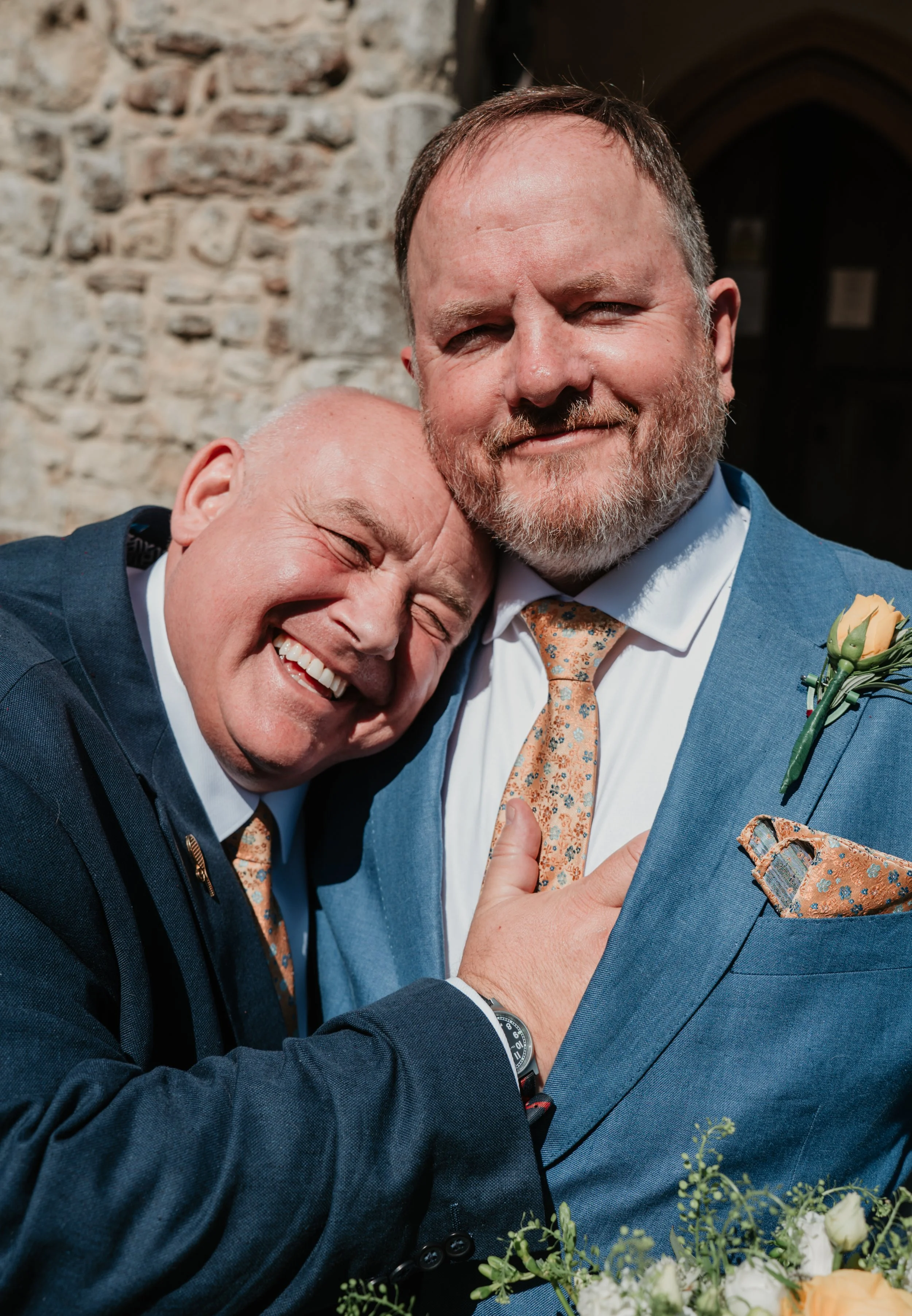 Two men in suits embracing and smiling, with one man resting his head on the other's shoulder. The man on the right wears a blue suit with a floral tie, and the man on the left wears a dark suit with a white shirt. They are outdoors against a stone w