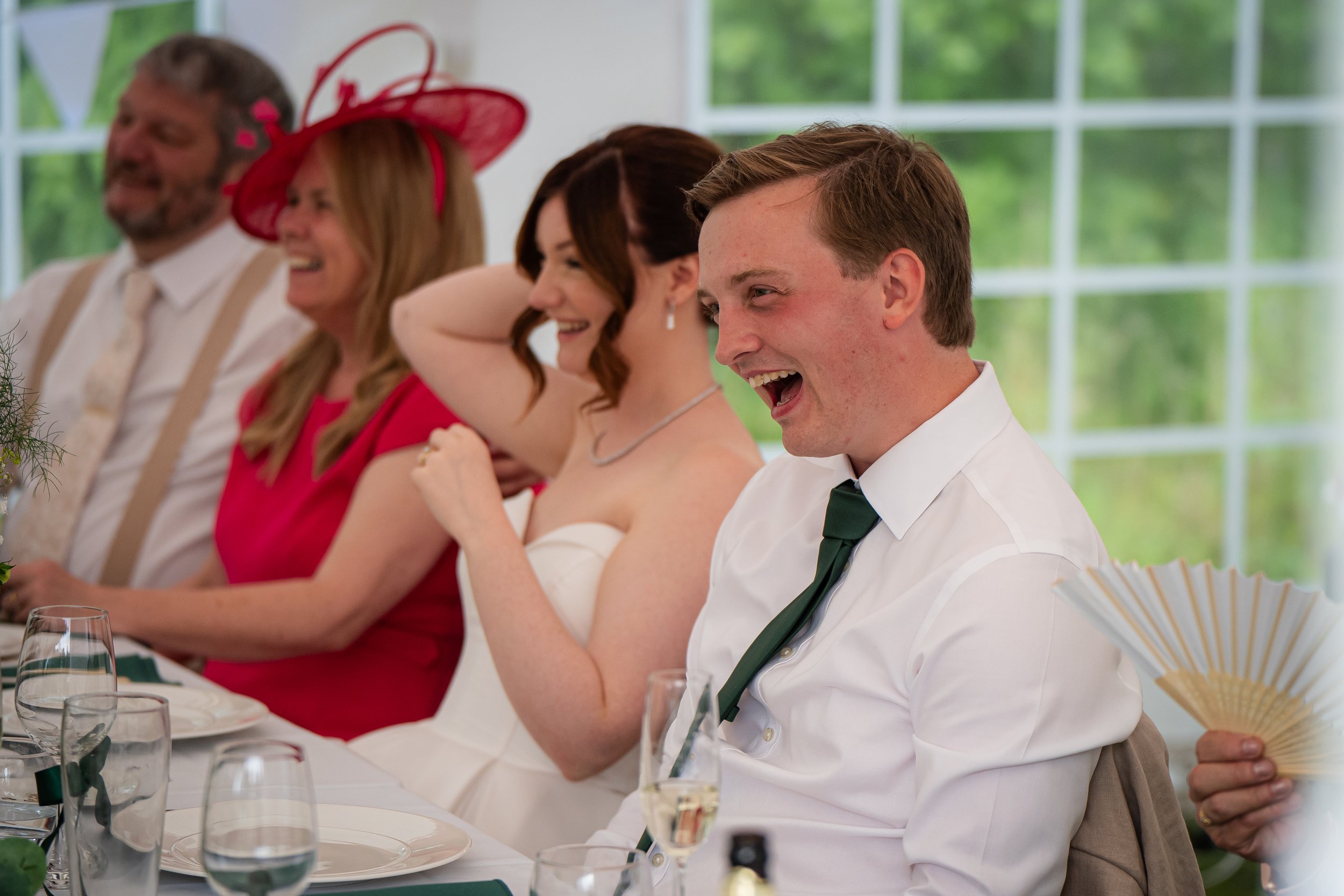People sitting at a table, smiling and laughing, during a social gathering or celebration in a well-lit room with large windows showing greenery outside.